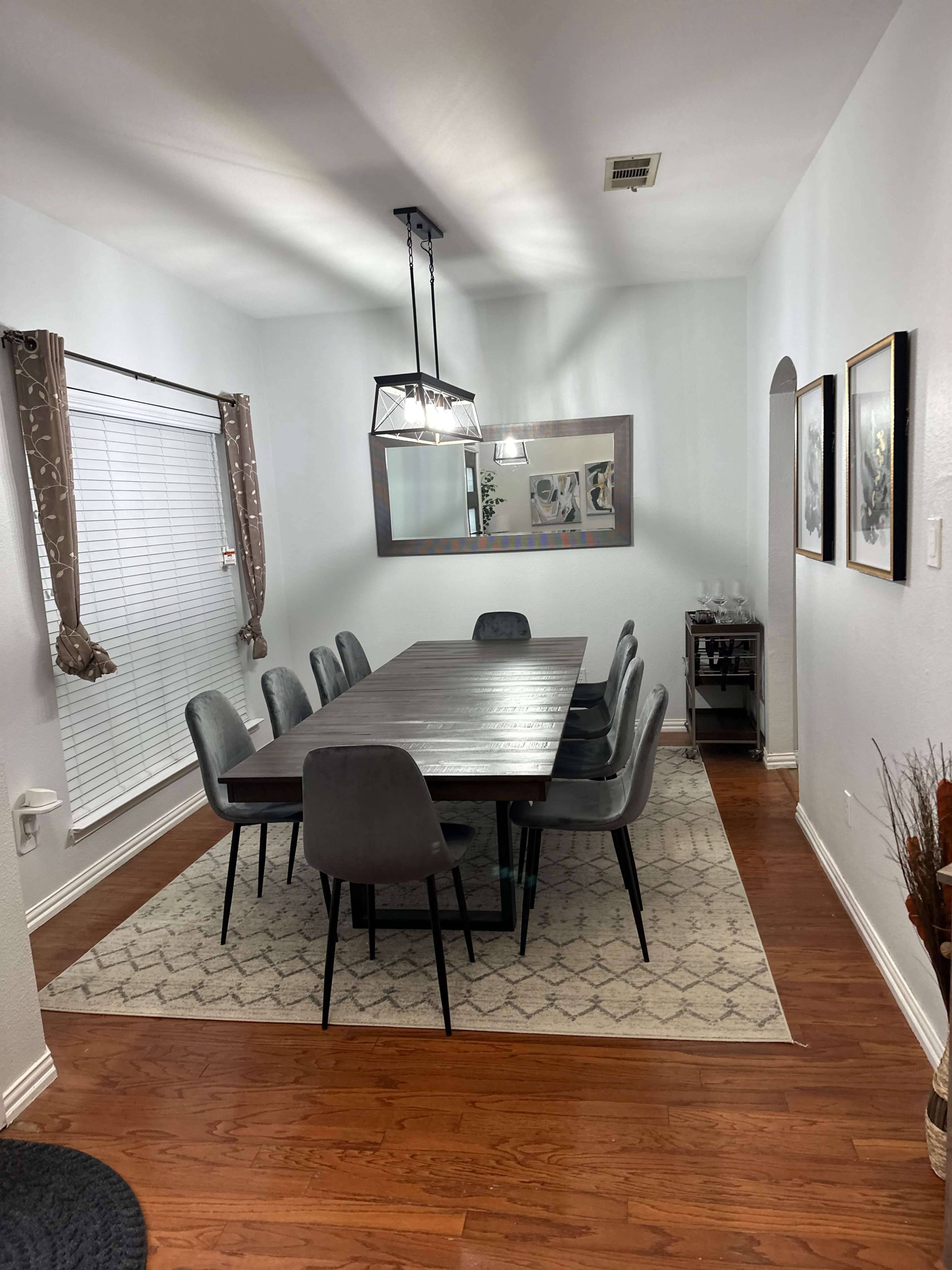 A modern dining room features a large wooden table surrounded by eight gray chairs, complemented by a geometric rug, pendant lights, and decorative mirrors on the walls.