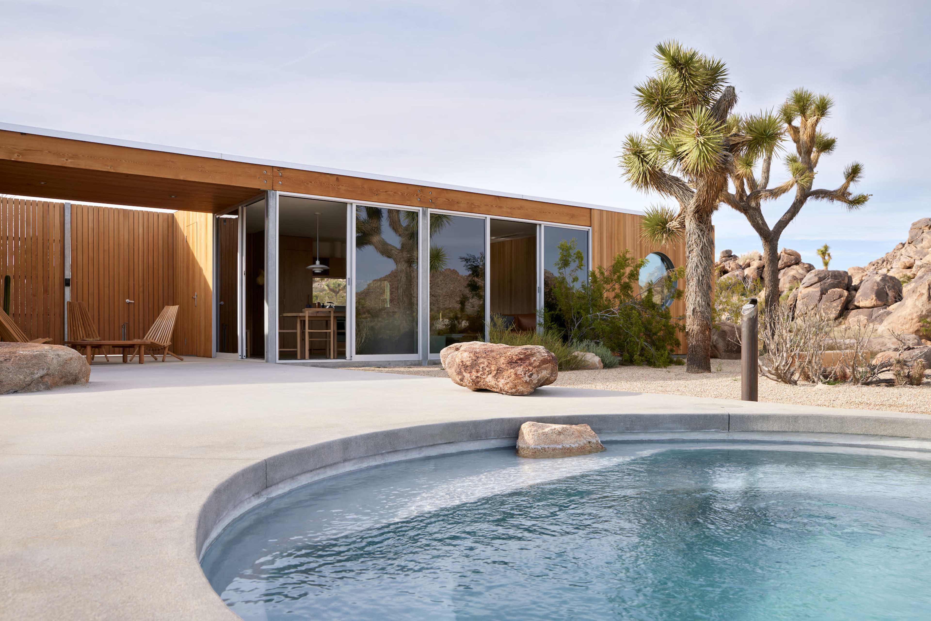 A modern, geometric home with large glass doors opens to a circular pool, surrounded by desert landscaping and rock formations.