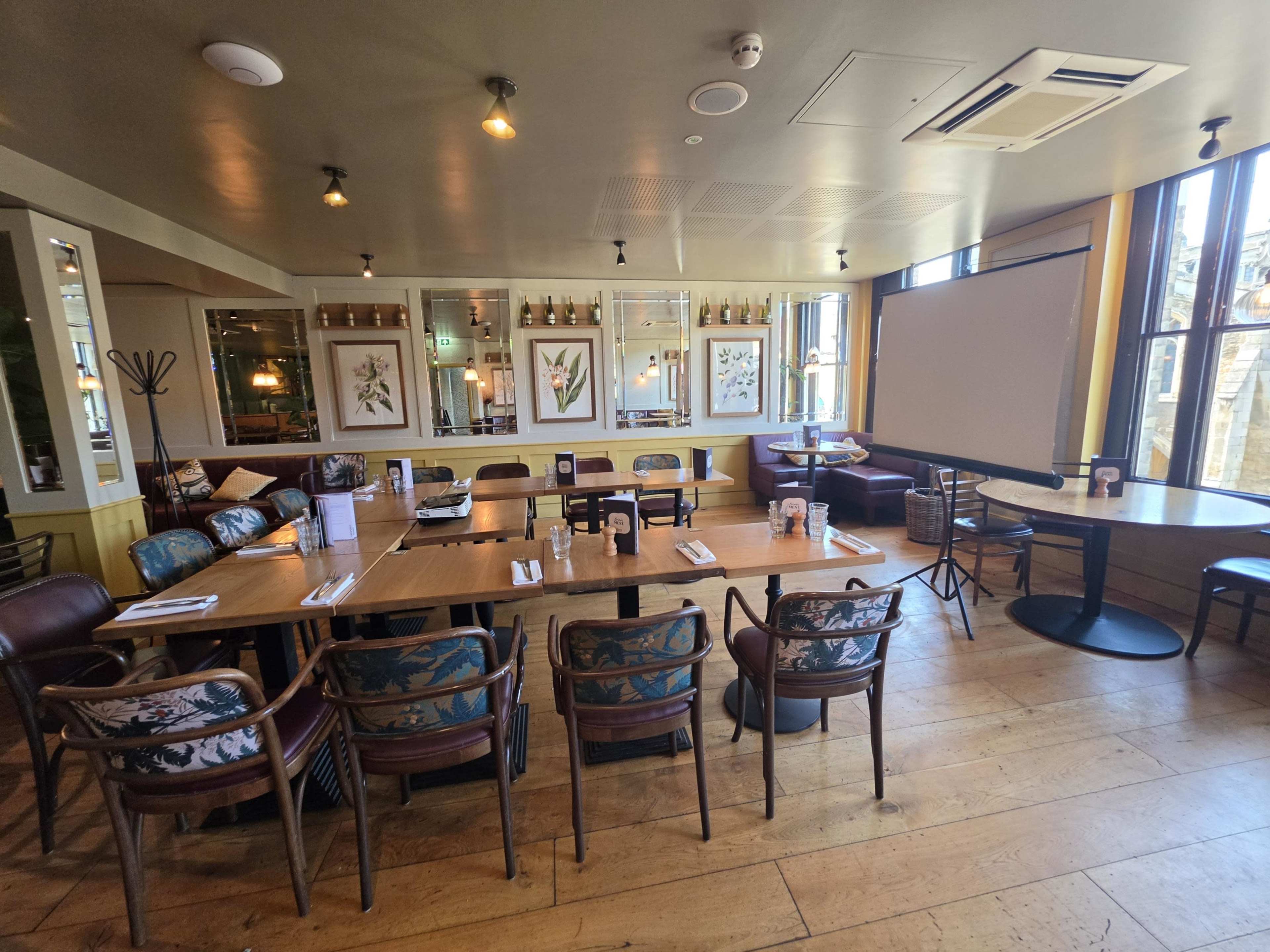 The interior of a restaurant features wooden tables, patterned chairs, and large windows showcasing natural light.