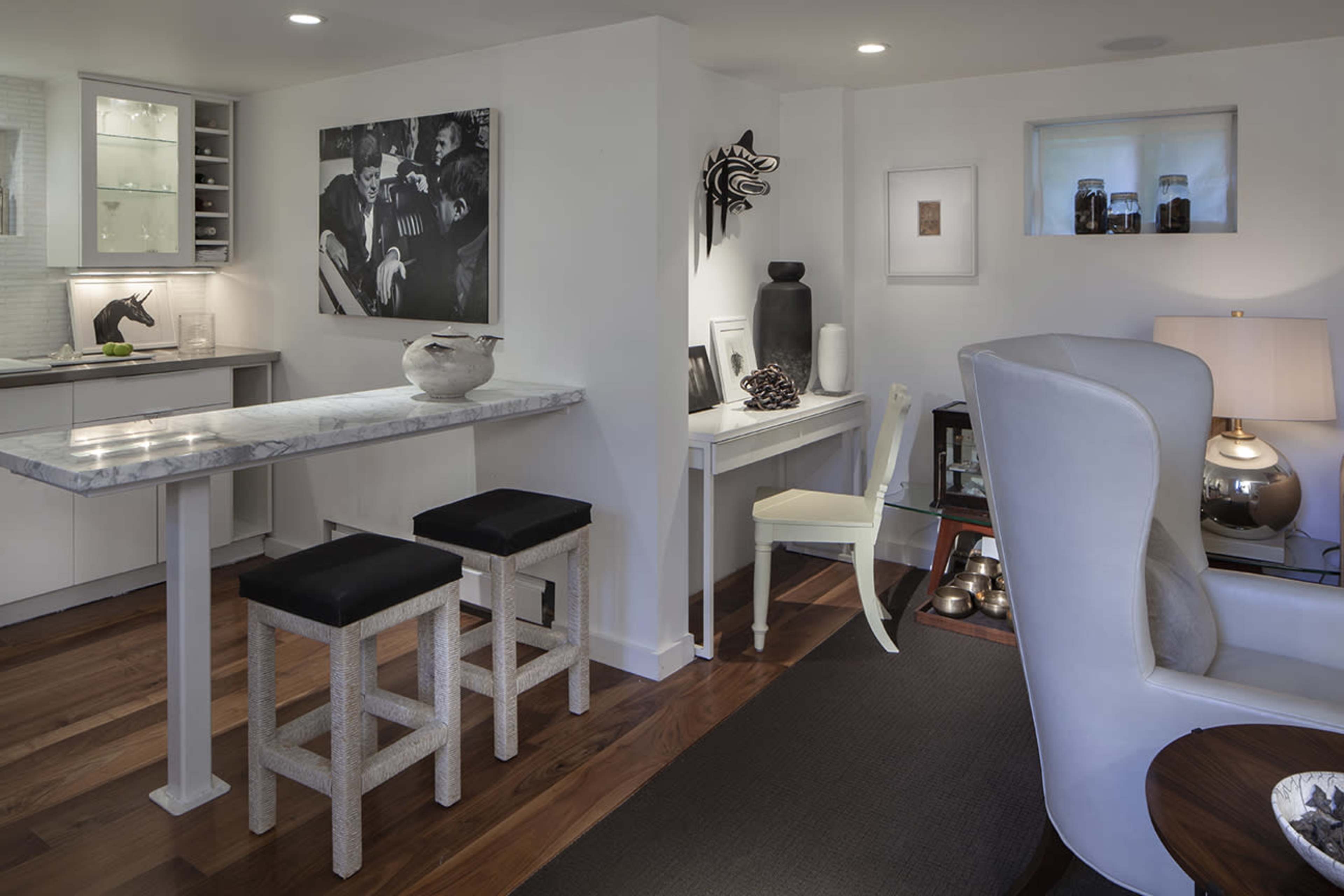 The image shows a modern indoor space featuring a small dining area with a marble-top table and two stools, alongside a minimalist desk, a stylish chair, and artwork on the walls.