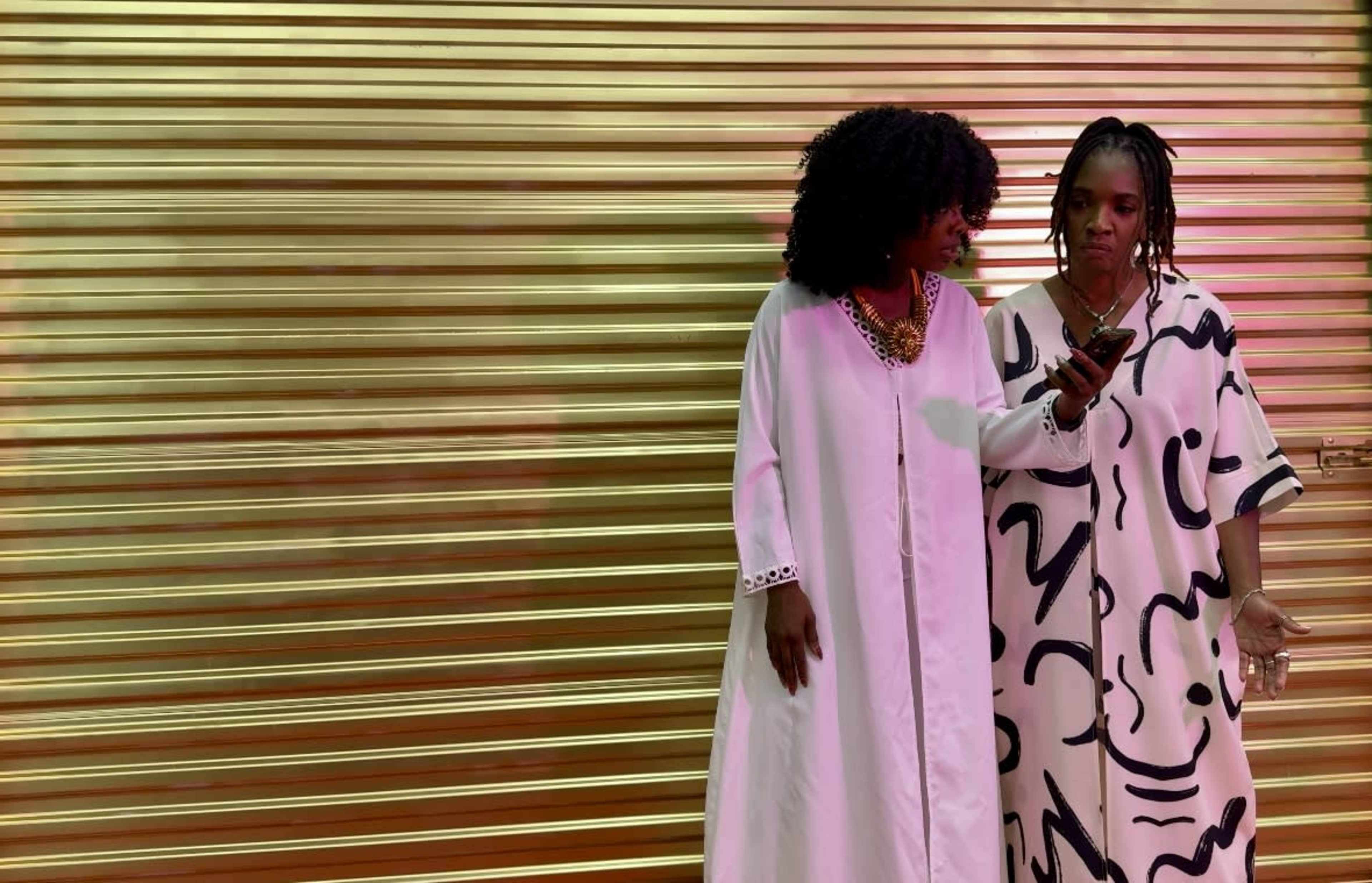 Two women stand side by side in front of a golden shutter, one checking her phone while the other looks on.