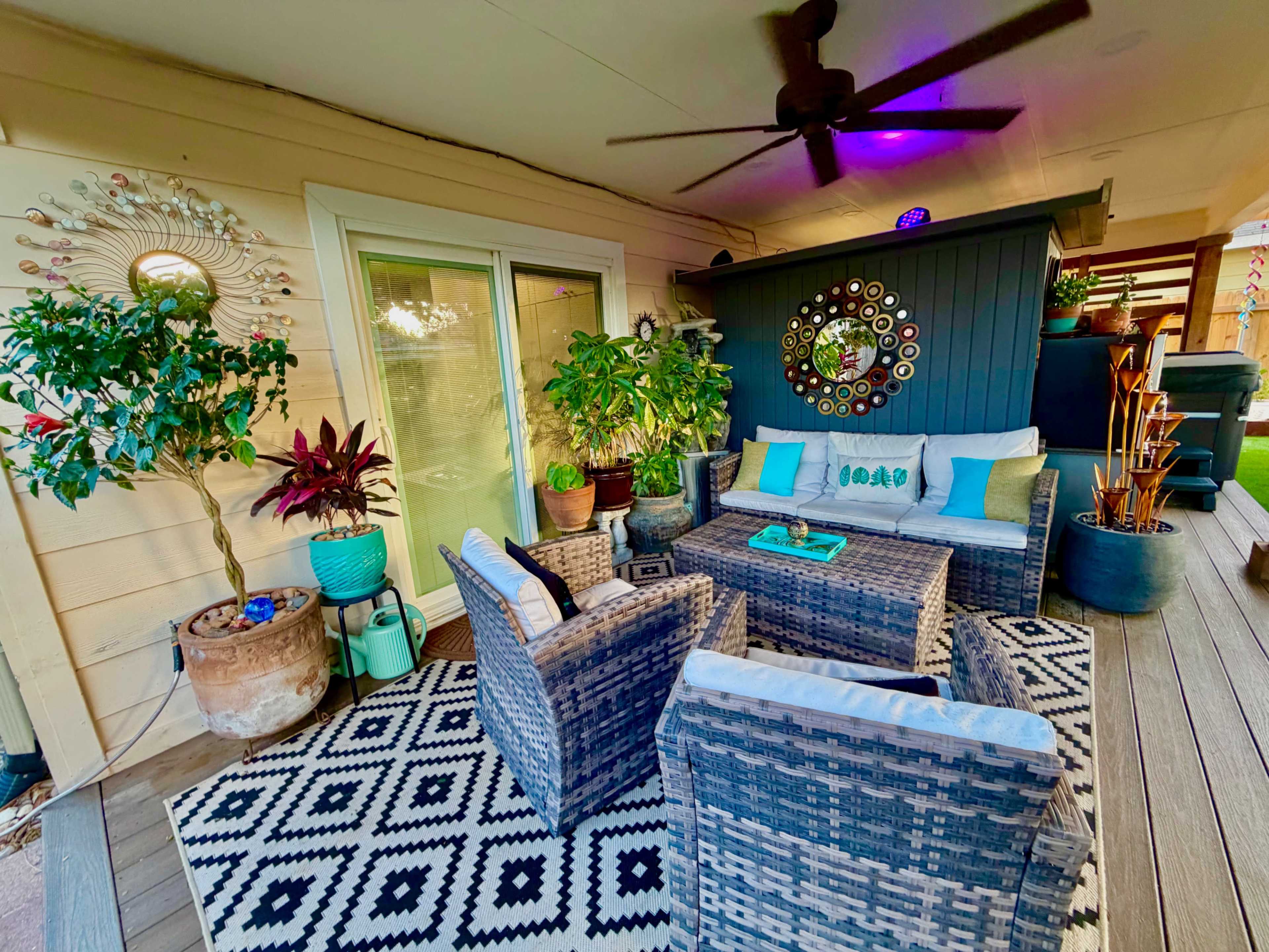 The image shows a cozy outdoor seating area with wicker chairs, green plants, and decorative wall art, all arranged on a patterned rug under a ceiling fan.