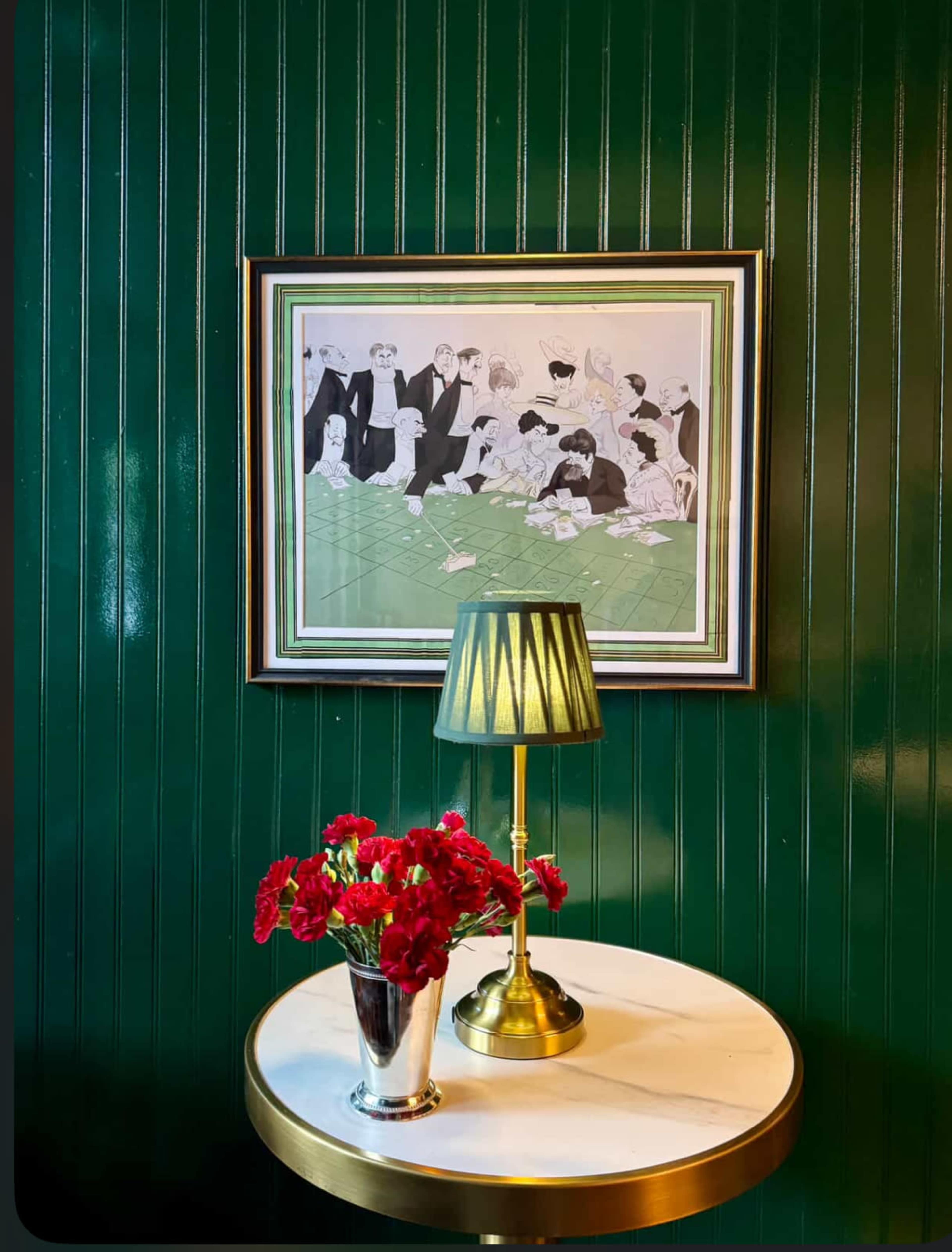 A green-paneled wall displays a framed artwork above a round table topped with a vase of red flowers and a gold-accented lamp.