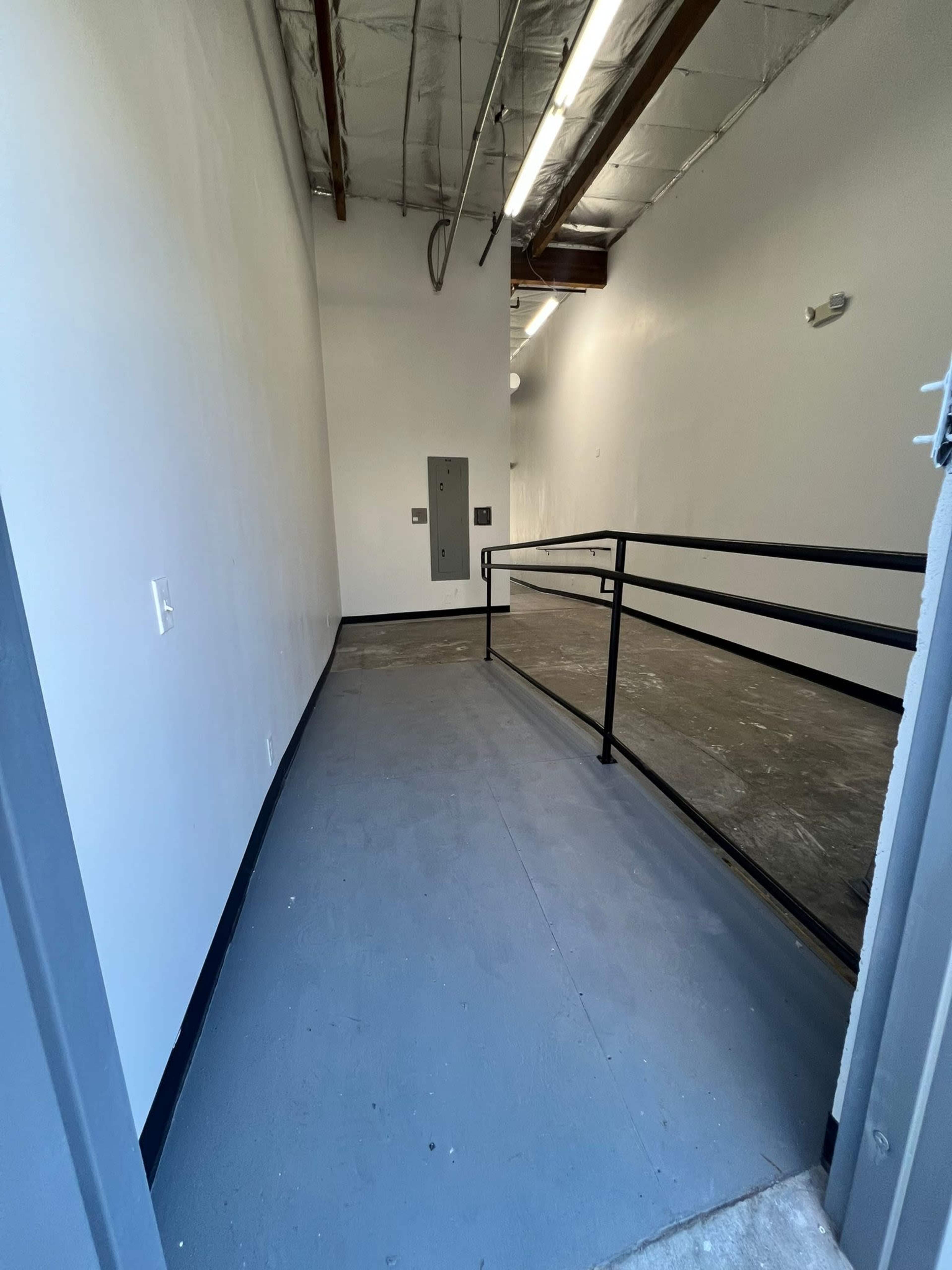 The image shows a wide, empty hallway with gray flooring and white walls, leading to a ramp and a utility panel.
