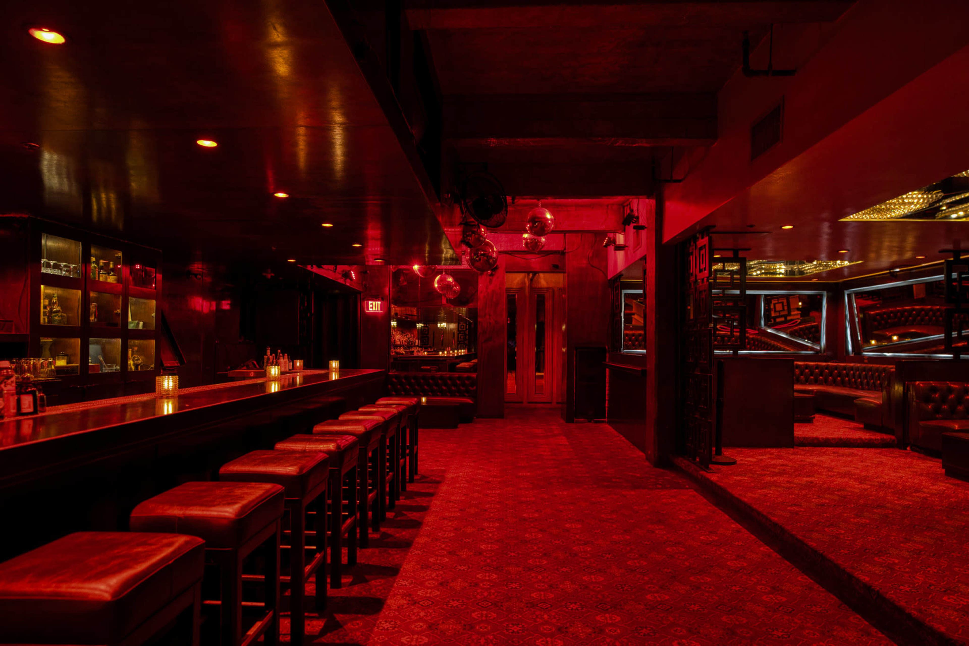 The interior of a dimly lit bar features a long counter with high stools, red-tinted lighting, and plush seating areas.