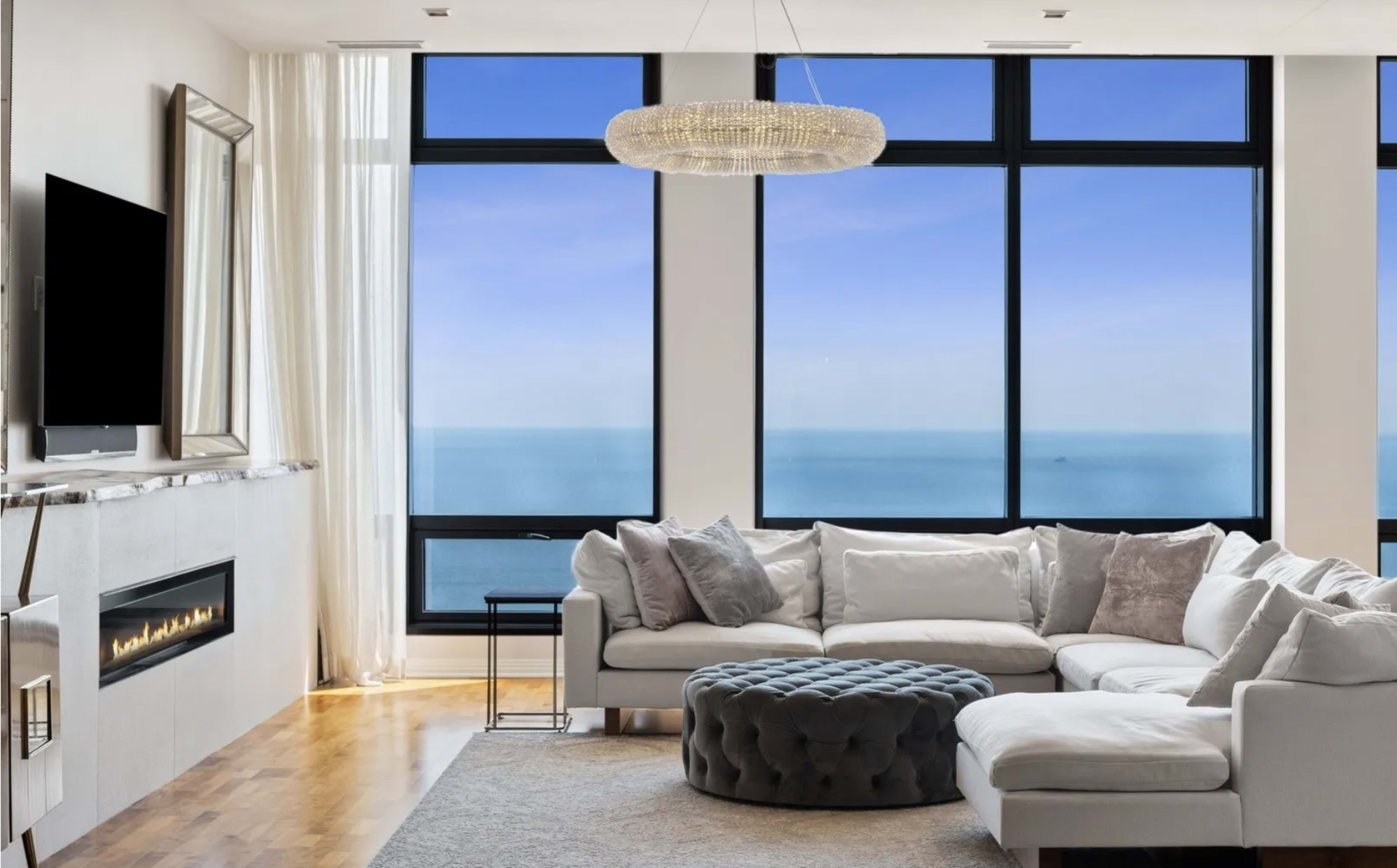 A modern living room features a sectional sofa, a round ottoman, and large windows offering a view of the ocean.