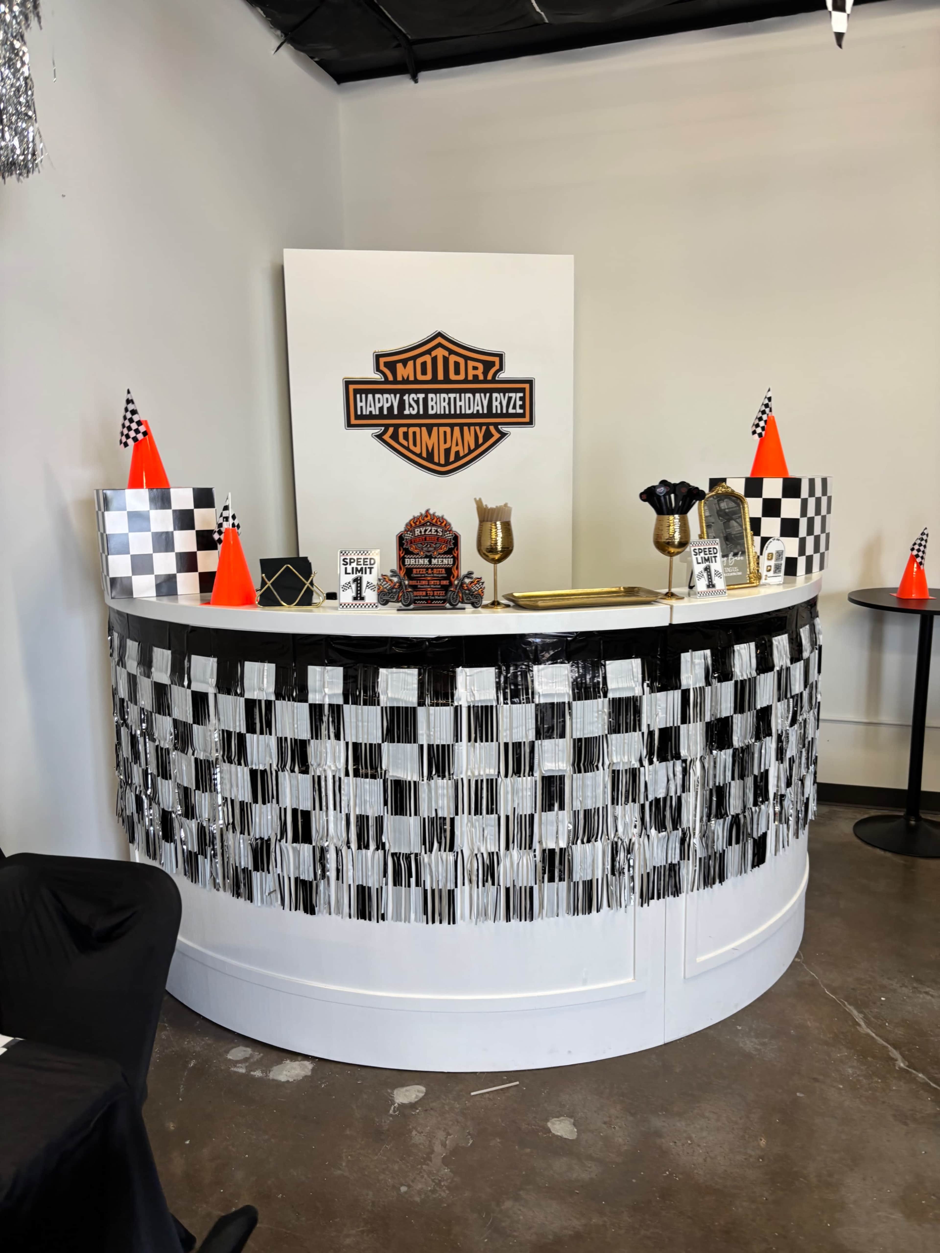 The image shows a decorated birthday party reception desk with a checkered flag design, various party supplies, and a sign reading "Happy 1st Birthday Ryze."