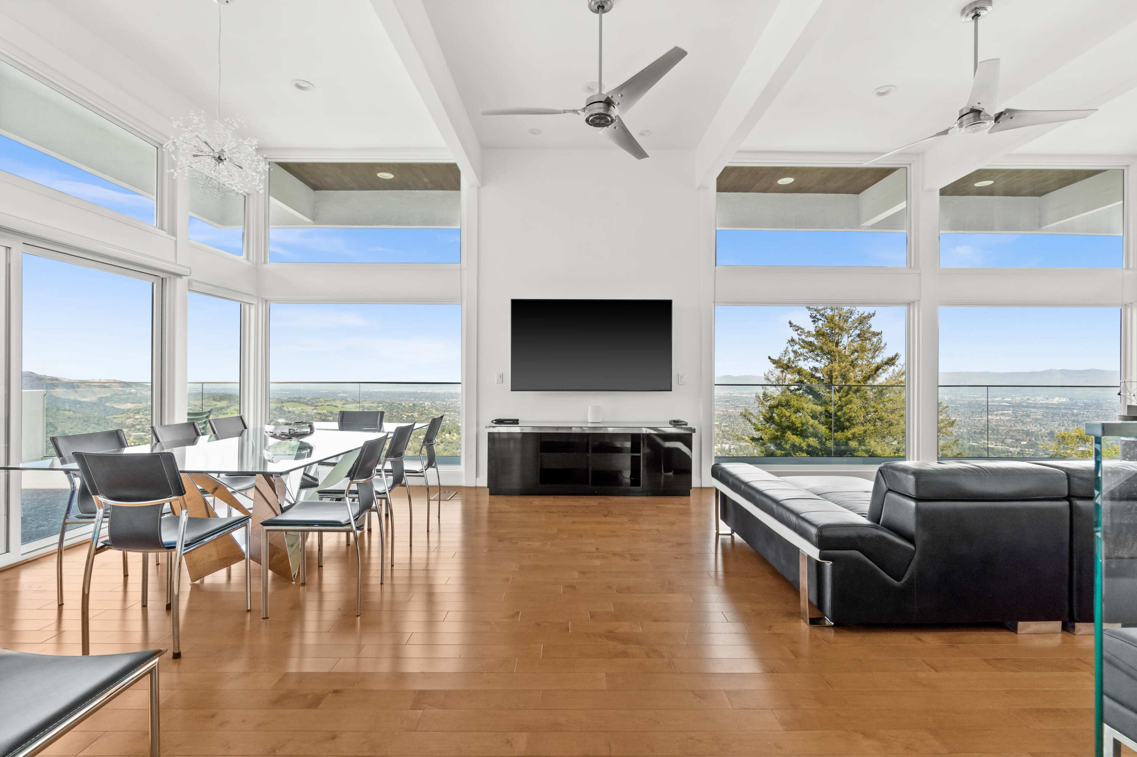 A modern living space features large windows with a view, a black sectional sofa, a dining area with a glass table, and a wall-mounted television.