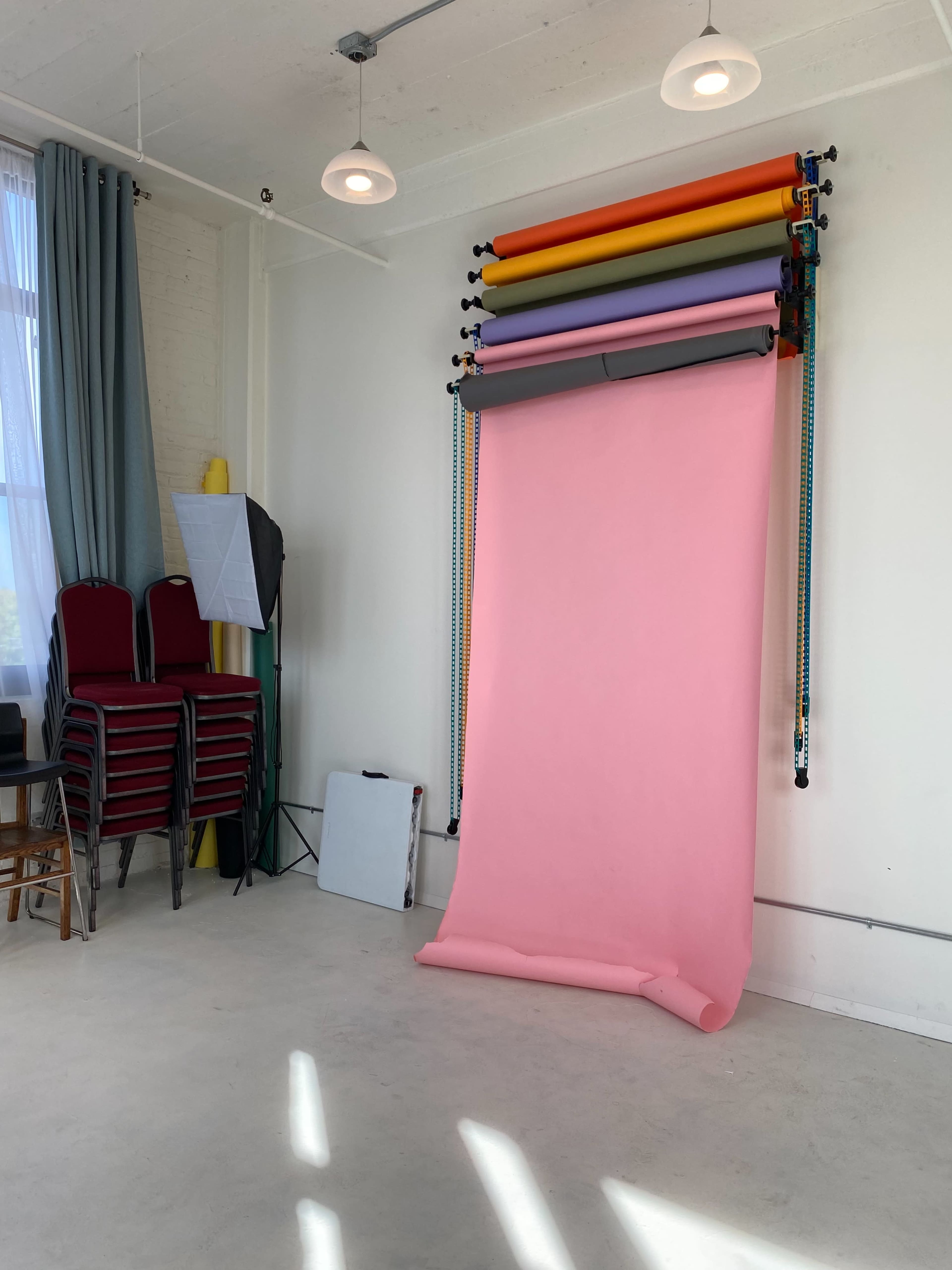 A photo studio with a pink backdrop setup against a white wall, alongside stacked chairs and various equipment.