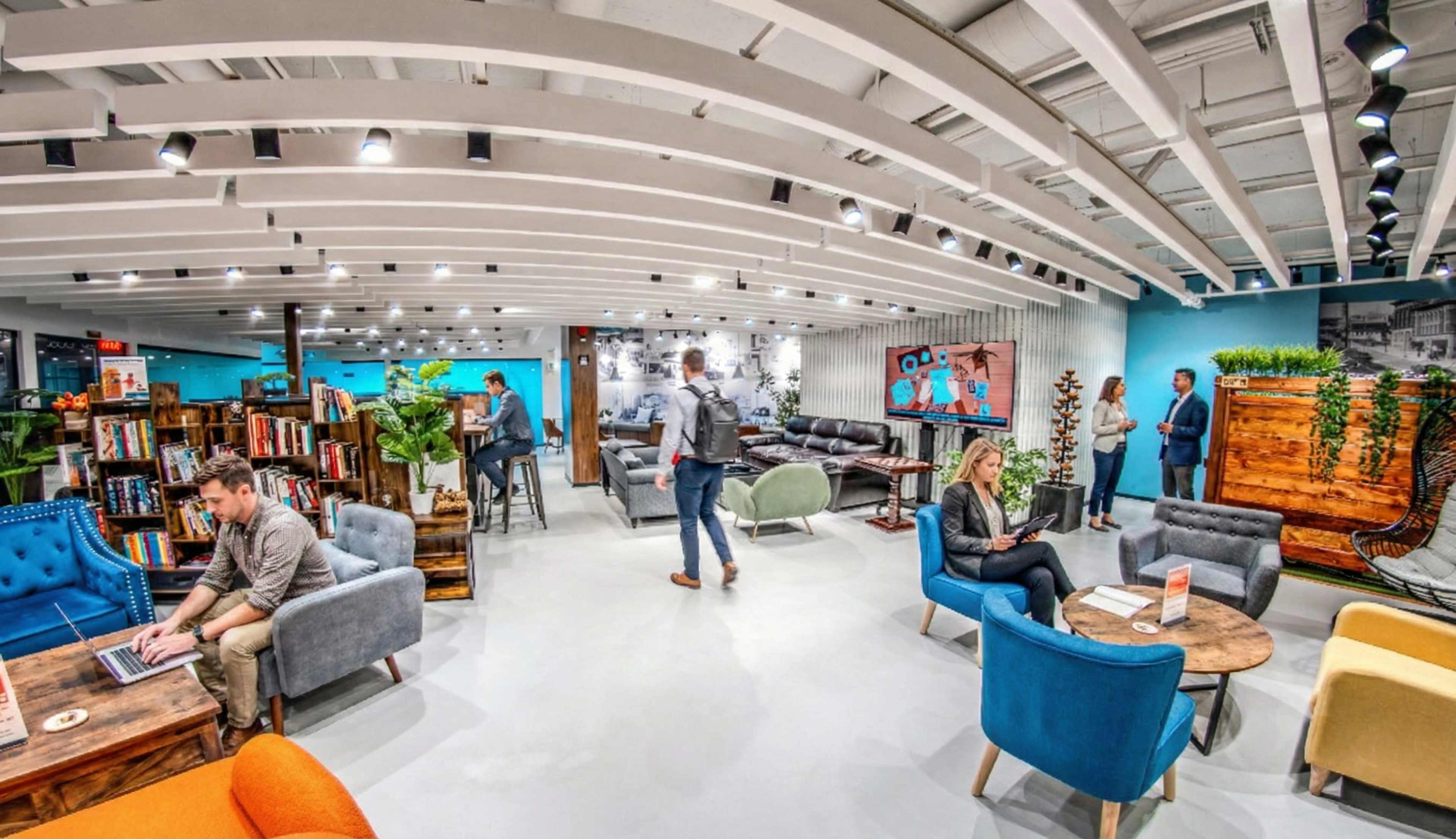 The image shows a modern office lounge area with comfortable seating, bookshelves, and people engaging in various activities.