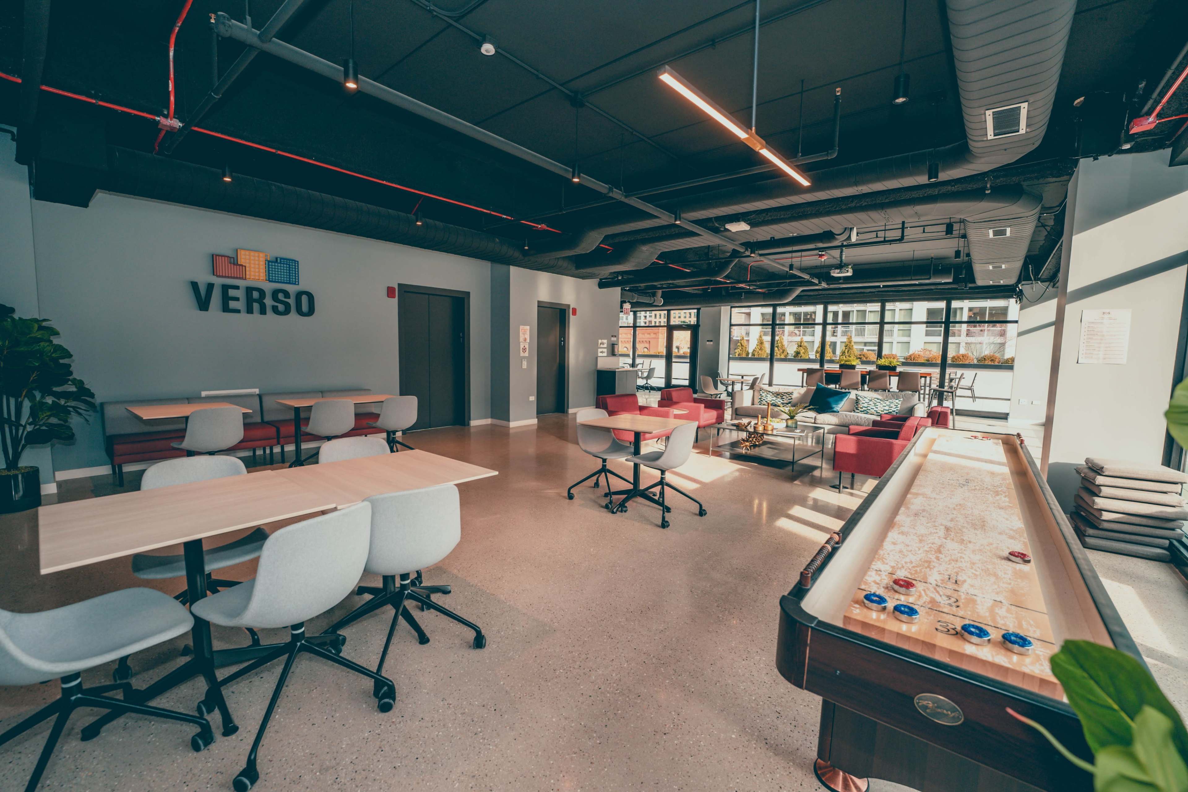 A modern communal workspace featuring tables, chairs, and a game area with a shuffleboard table, illuminated by natural light from large windows.
