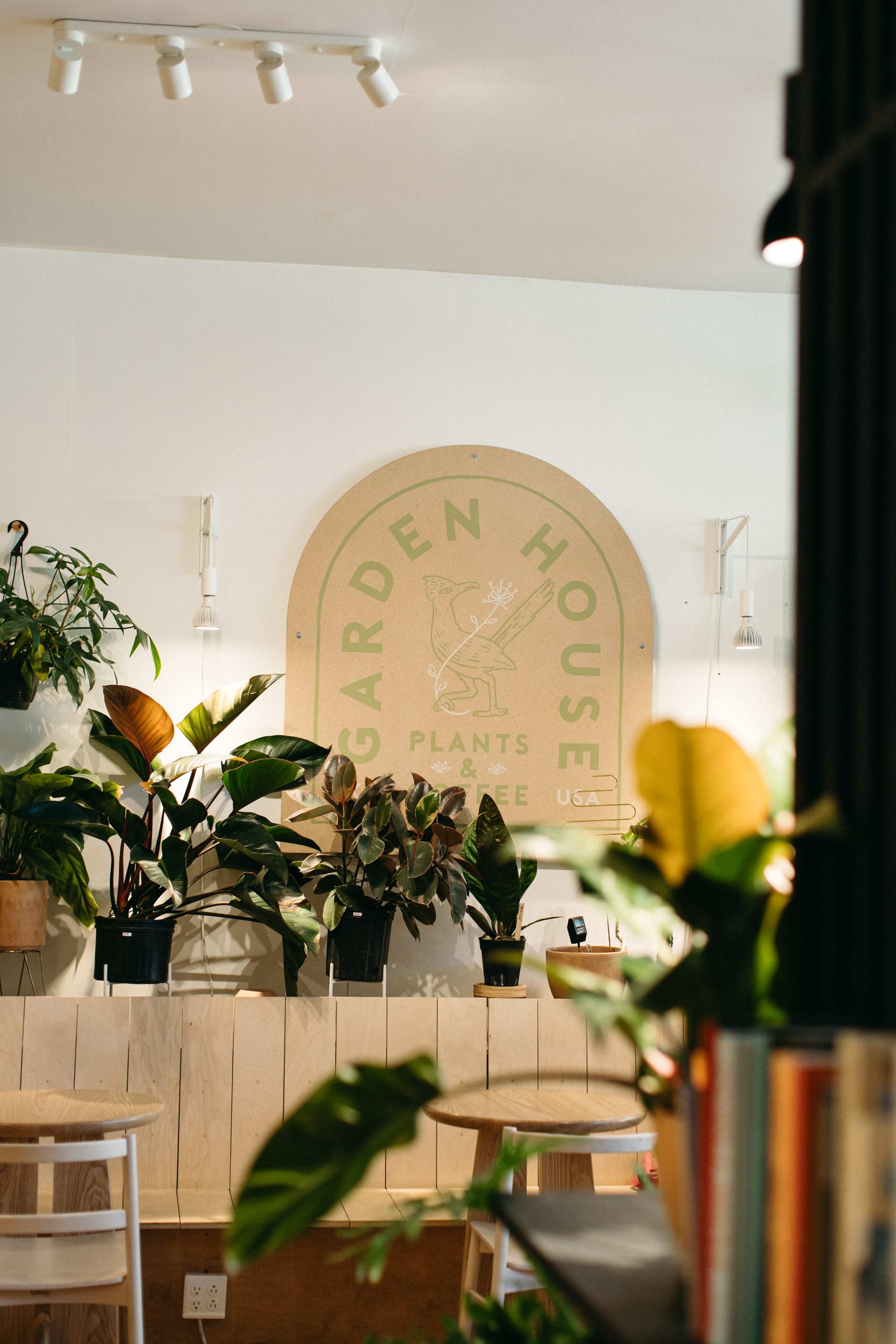 The image shows a cozy indoor space with a prominent sign reading "GARDEN HOUSE," surrounded by various potted plants and simple wooden furniture.