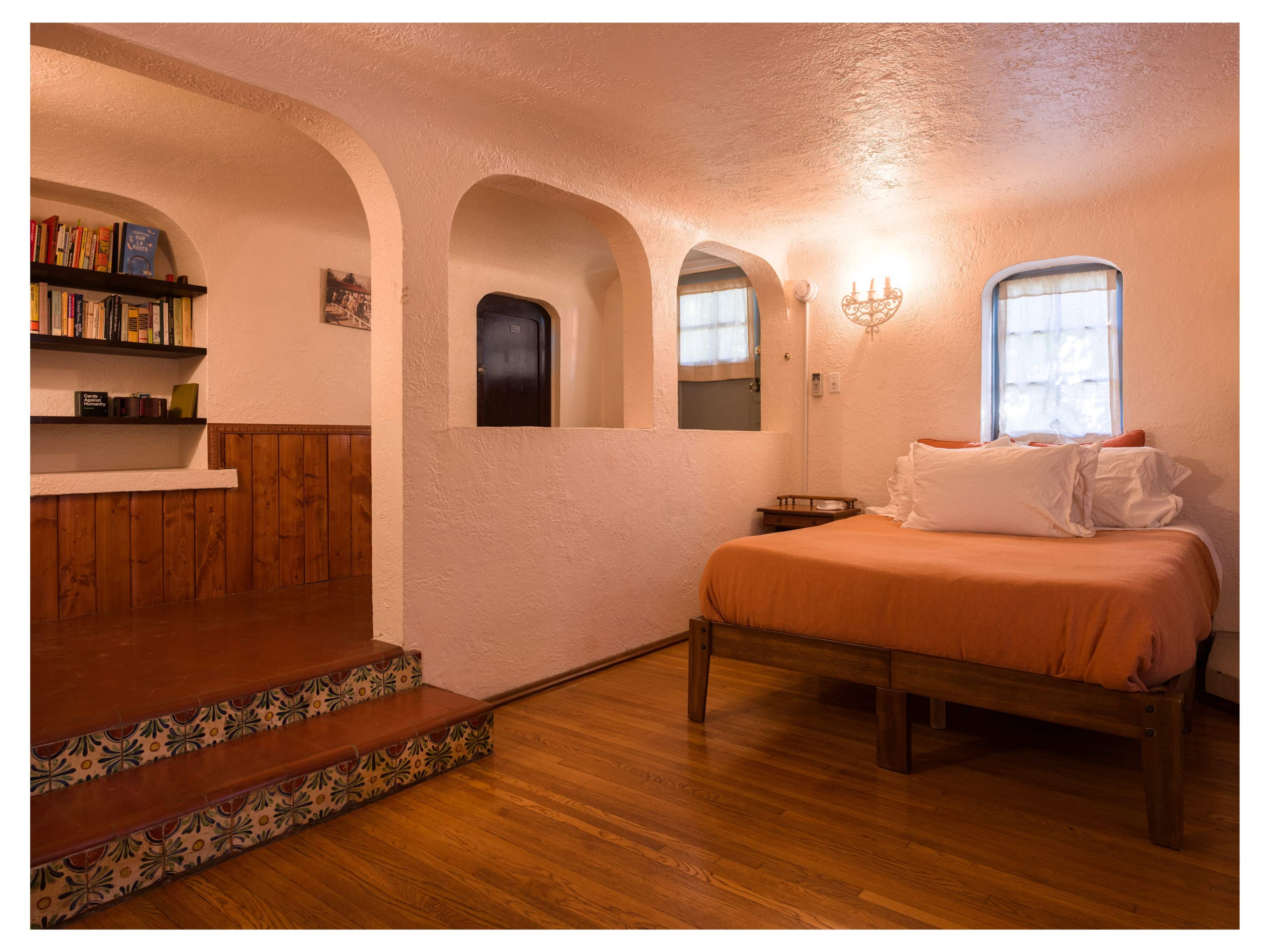 The image shows a cozy bedroom with a wooden bed, white walls, arched openings, and steps leading to a small seating area with bookshelves.