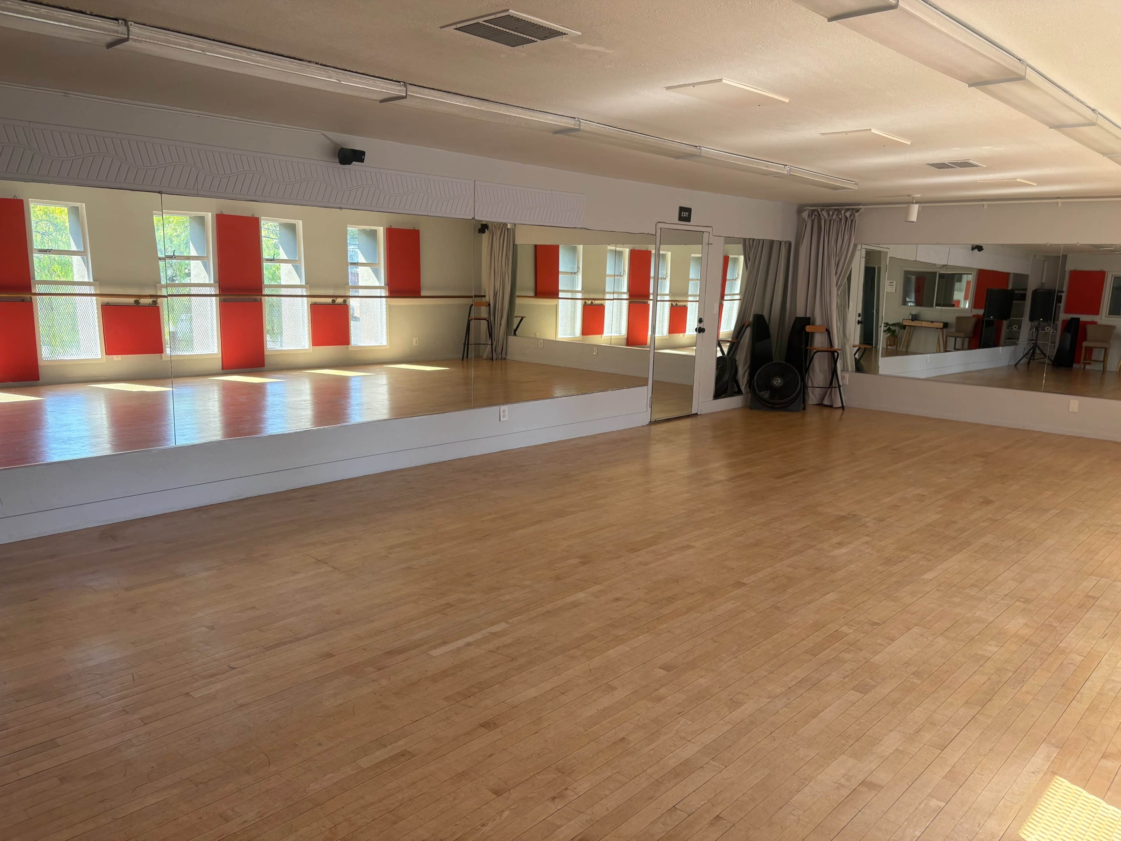 The image shows a spacious dance studio with wooden floors, large mirrors, and red accent walls.