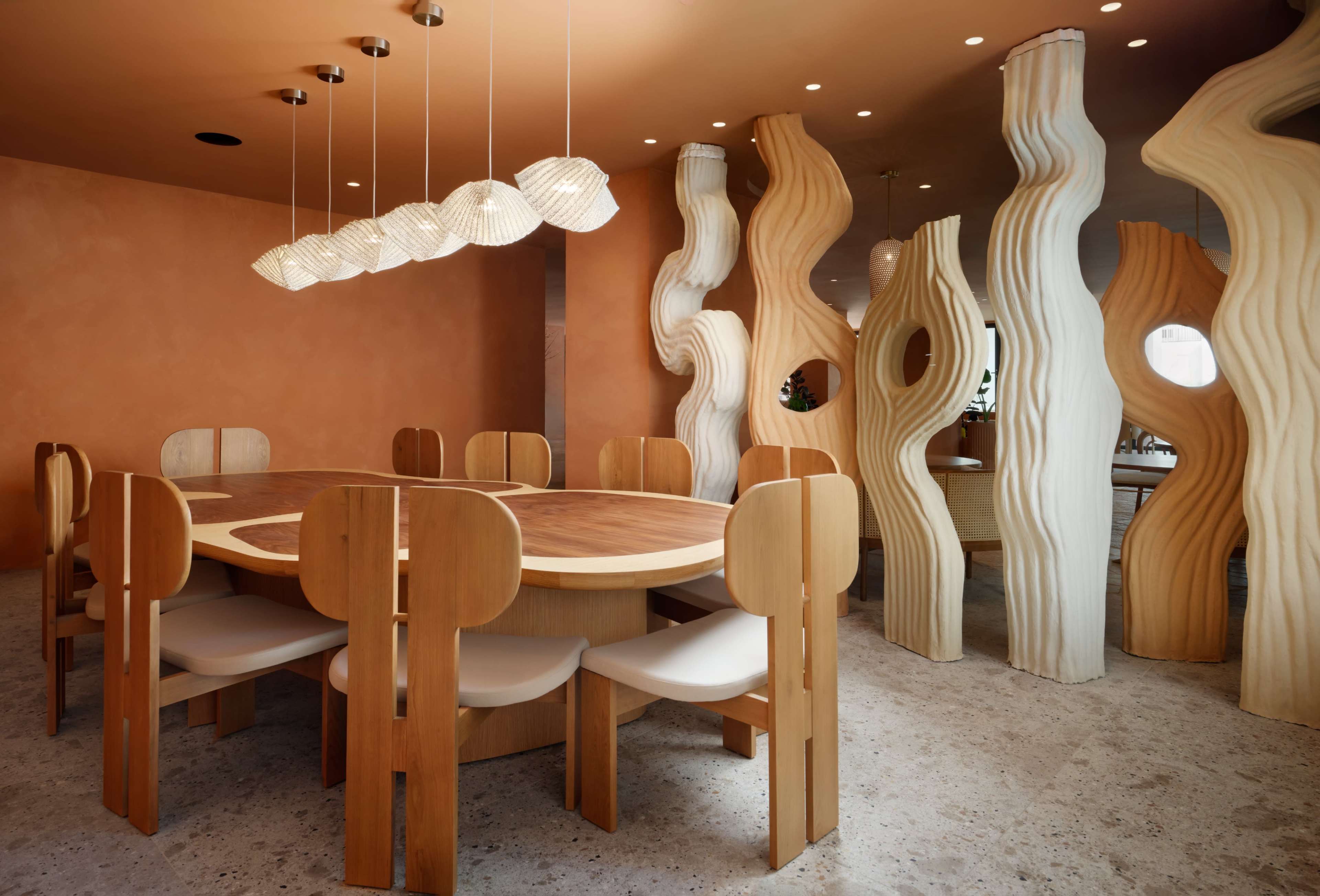 A modern dining area features a round wooden table surrounded by unique, sculptural wooden chairs and abstract column-like structures with circular cutouts.