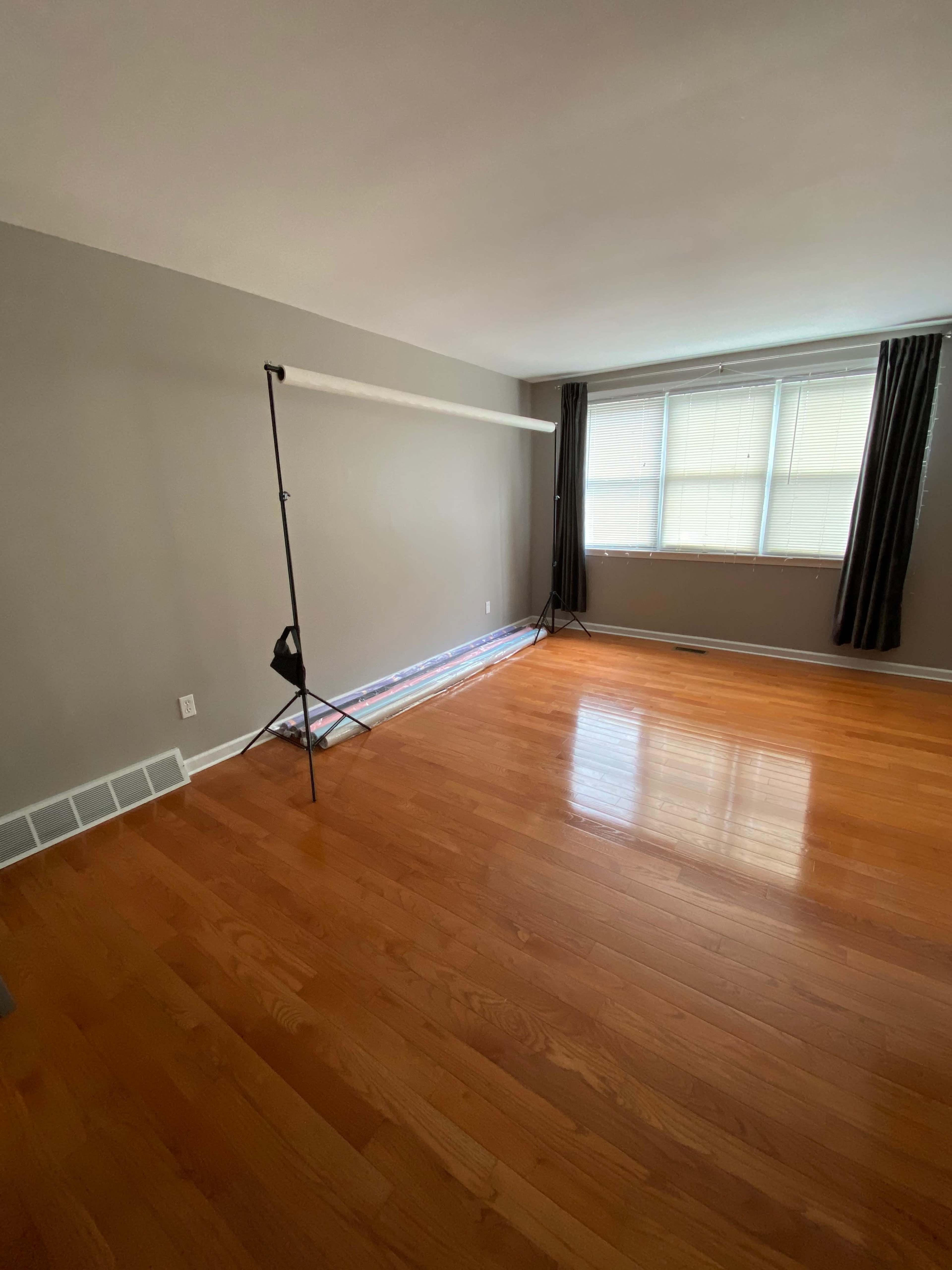 A spacious, empty room features polished hardwood floors, a large window with light-colored curtains, and a backdrop stand set up against one wall.
