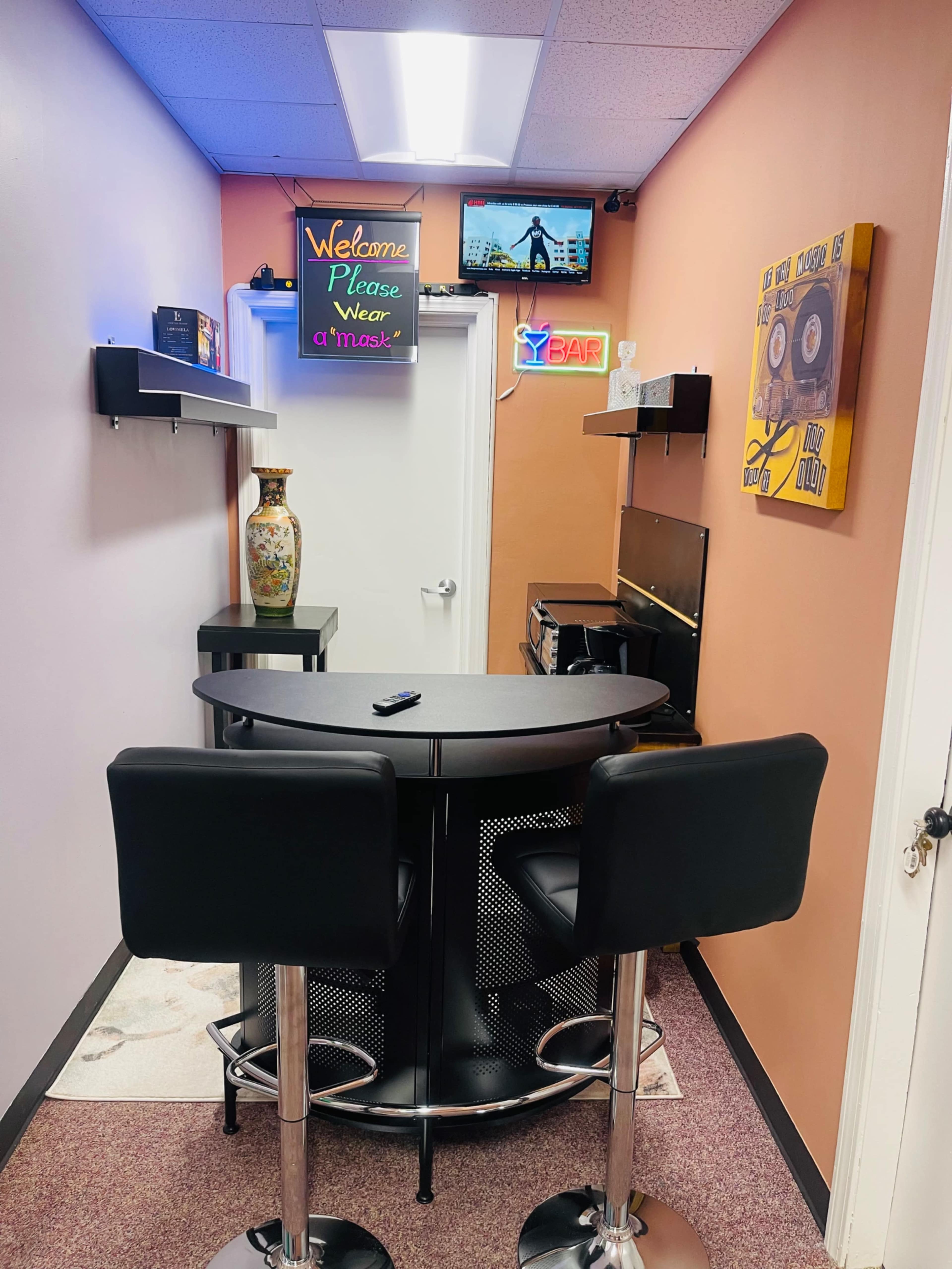 A narrow hallway with a round black table and two stools, decorated with a vase, wall art, and a television mounted on the wall.