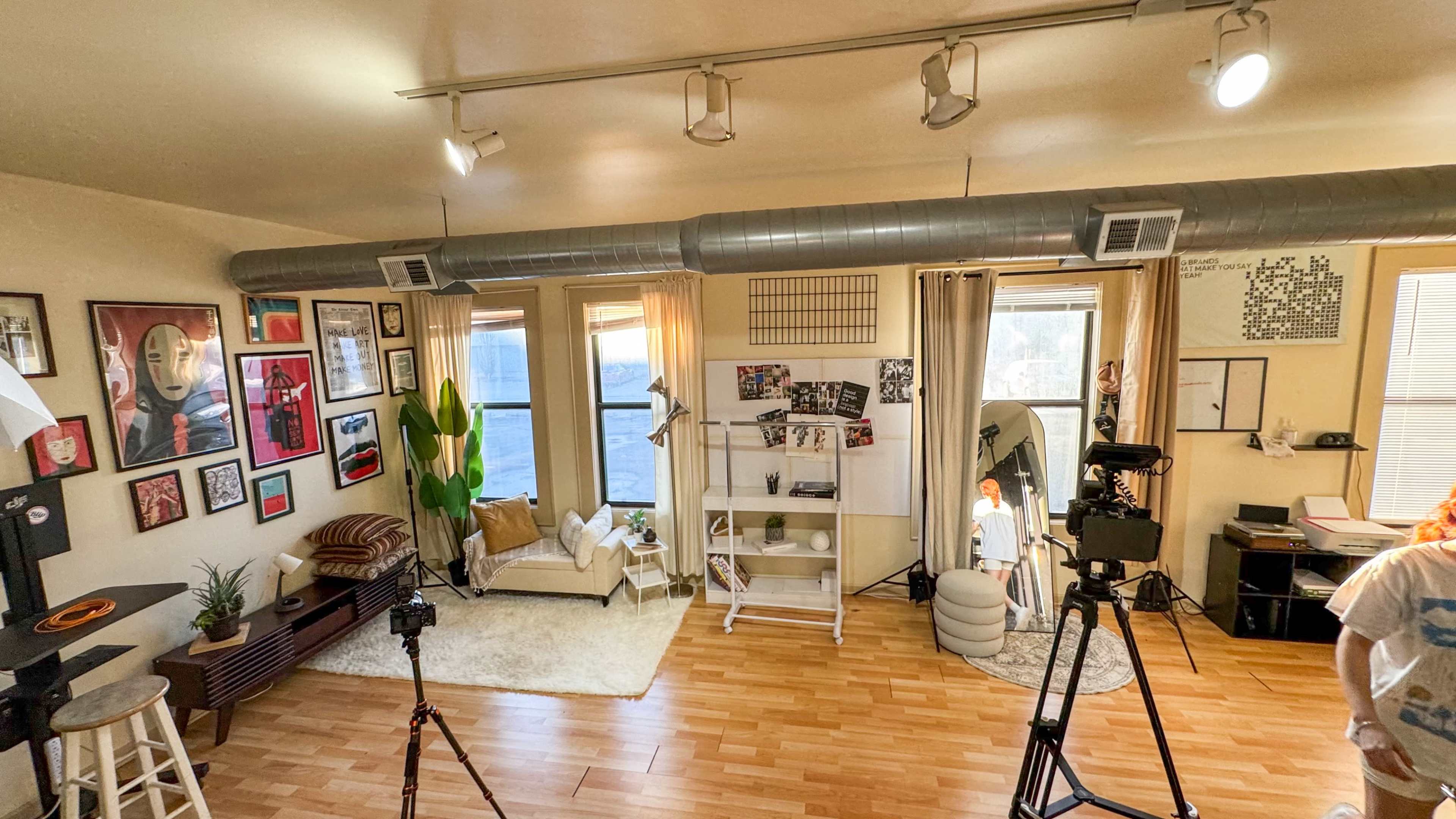 The image shows a well-lit room featuring a seating area with a couch and decorative plants, surrounded by framed artwork on the walls and two cameras positioned on tripods.