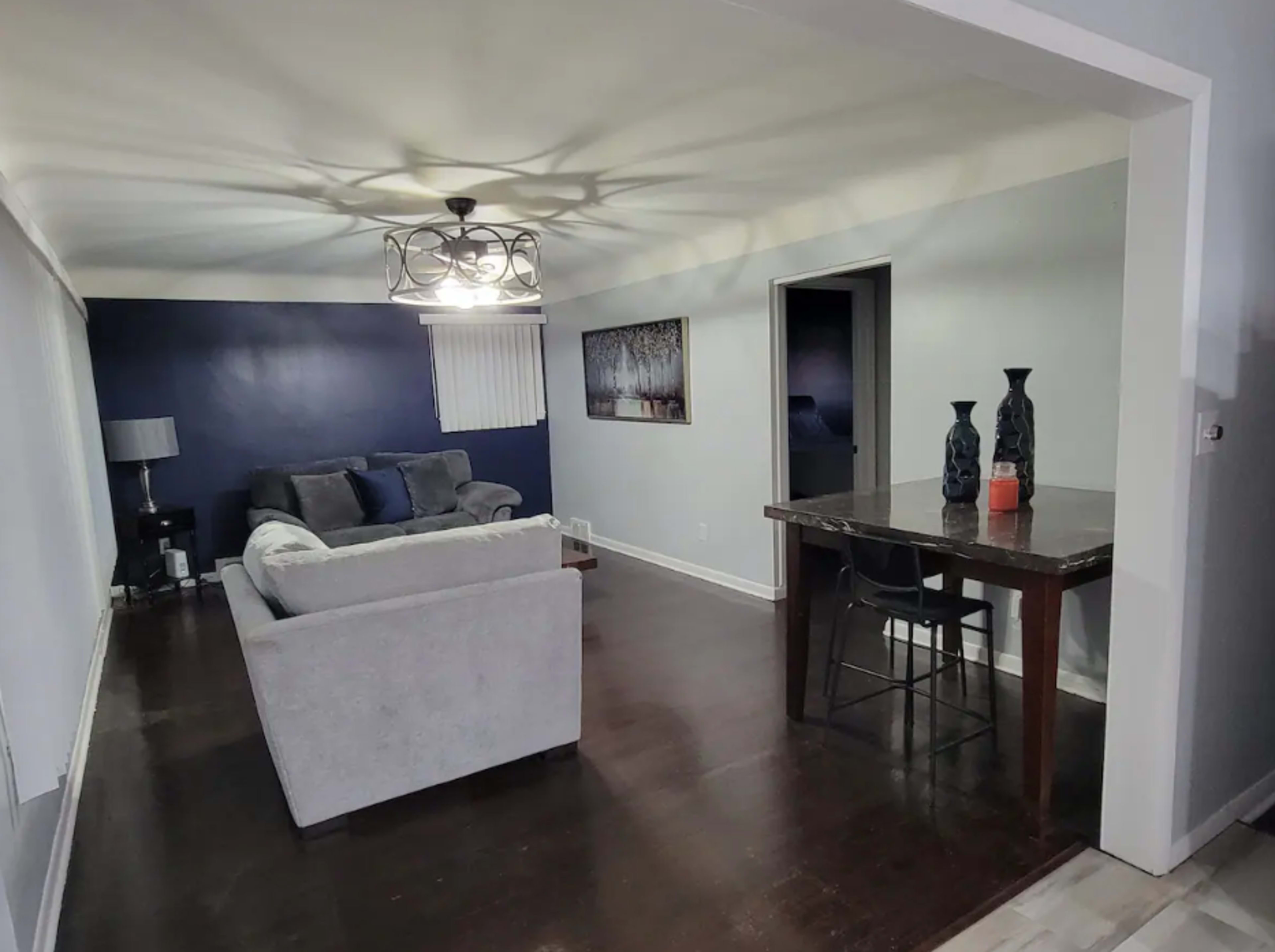 The image shows a living room with a gray sofa and a dining table, featuring a chandelier and a dark blue accent wall.
