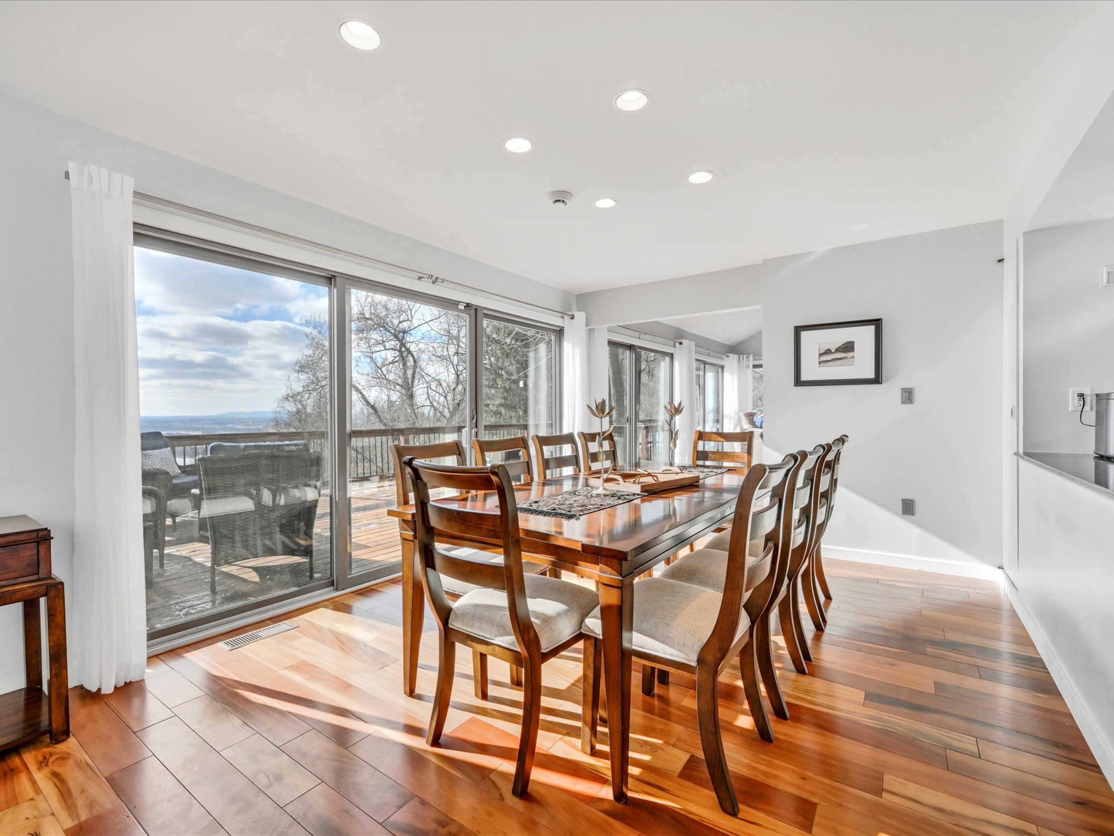 A spacious dining area features a large wooden table surrounded by chairs, with large windows offering a view of a deck and outdoor scenery.