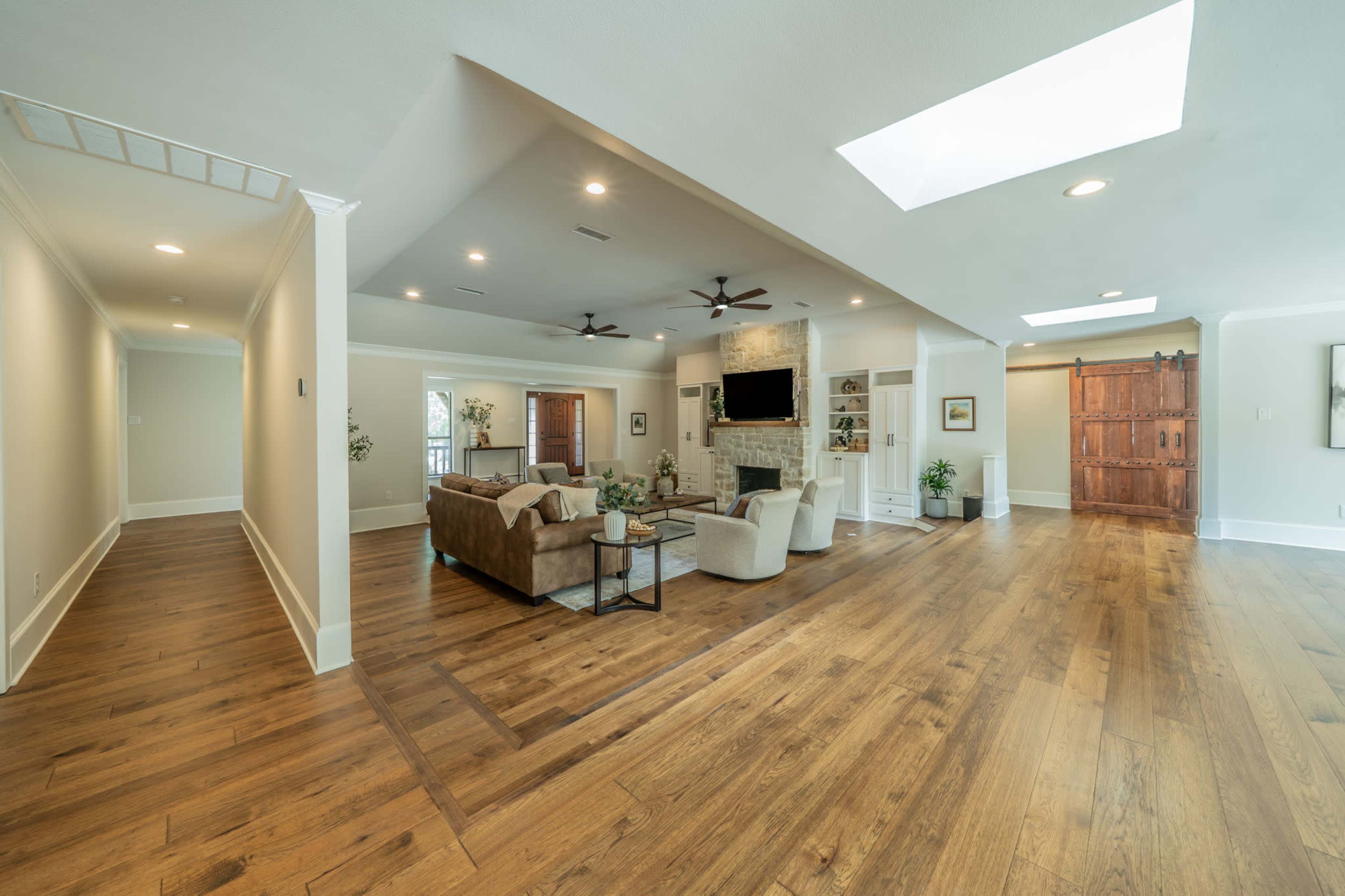 The image shows a spacious living room with hardwood flooring, a large sectional sofa, a mounted television, and natural light streaming through skylights.