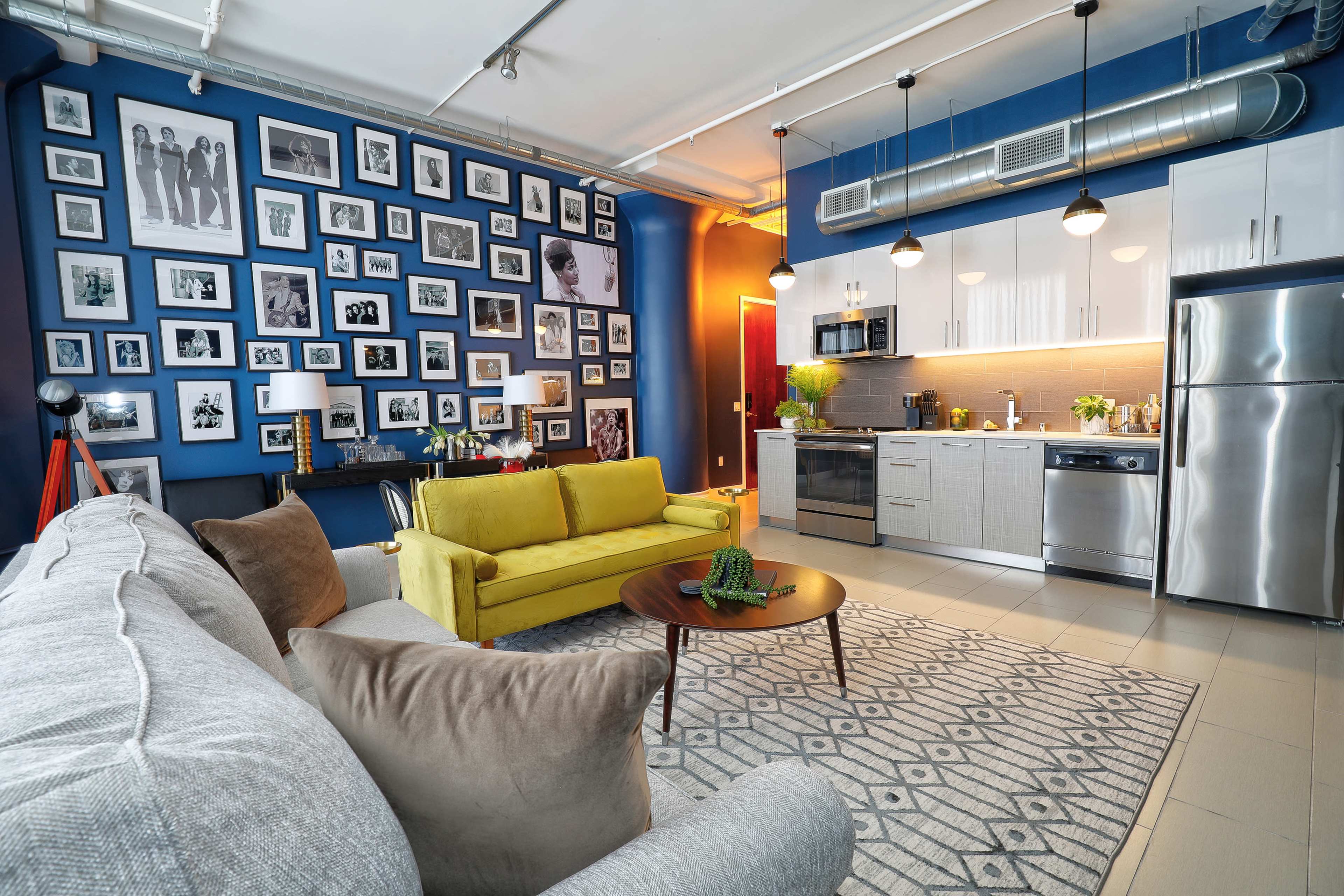 The image shows a modern living space with a yellow sofa, a gray couch, a round wooden coffee table, and a kitchen area featuring stainless steel appliances, all set against a blue wall adorned with a gallery of framed photos.