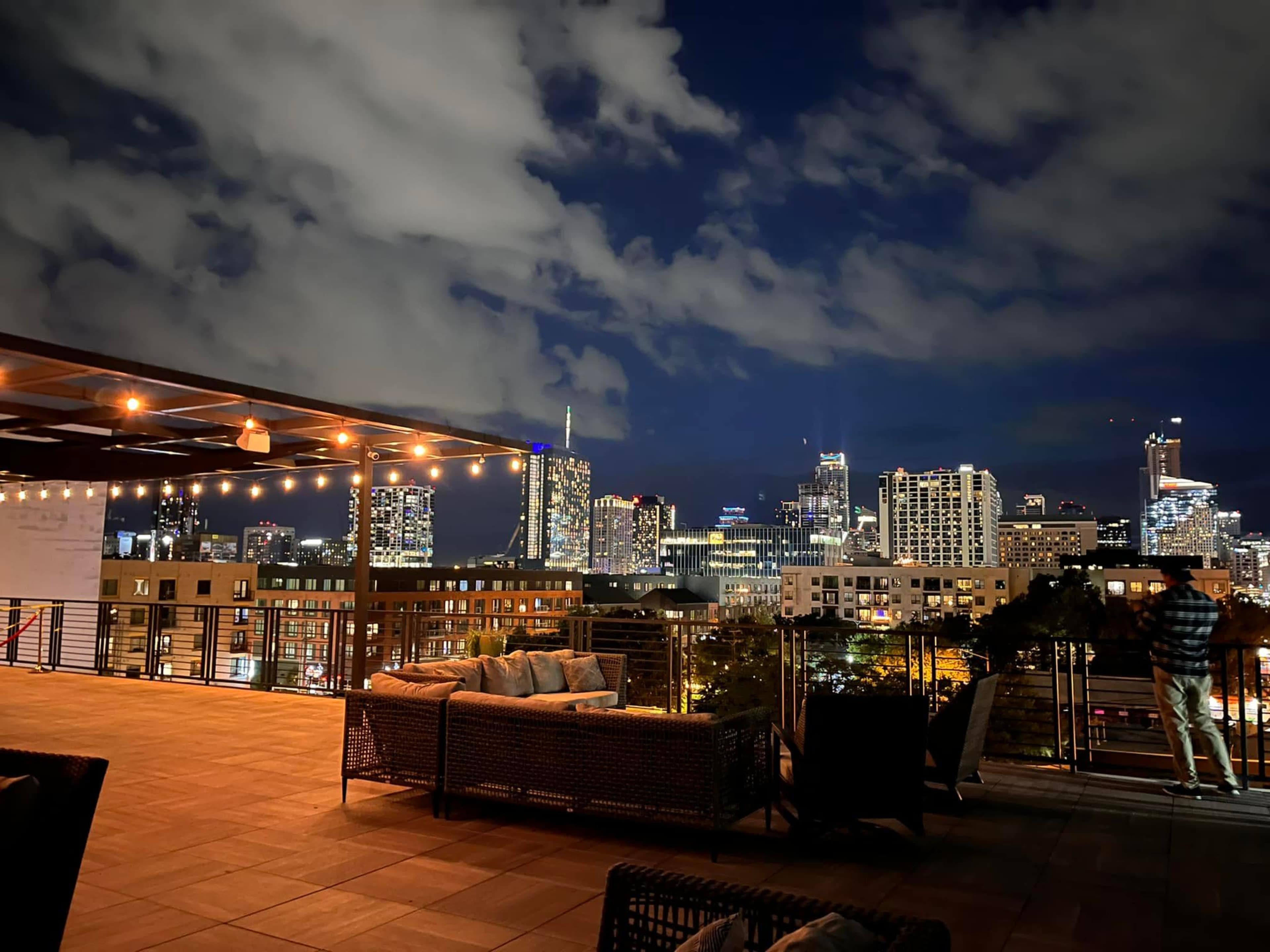 A rooftop patio features string lights and seating with a view of a city skyline at night.