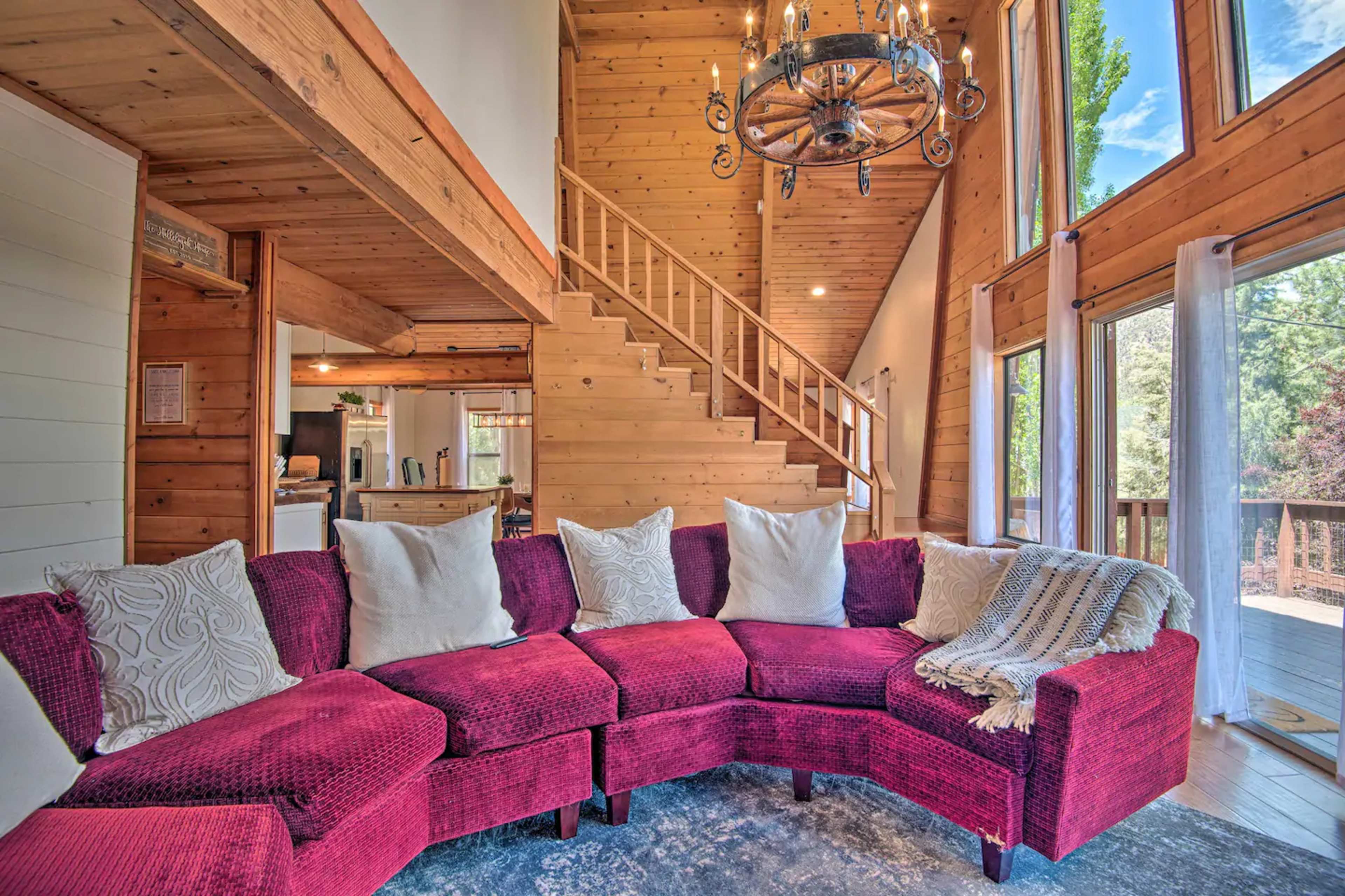A cozy living room features a red sectional sofa with white pillows and large windows, allowing natural light to illuminate the wooden interior and staircase.