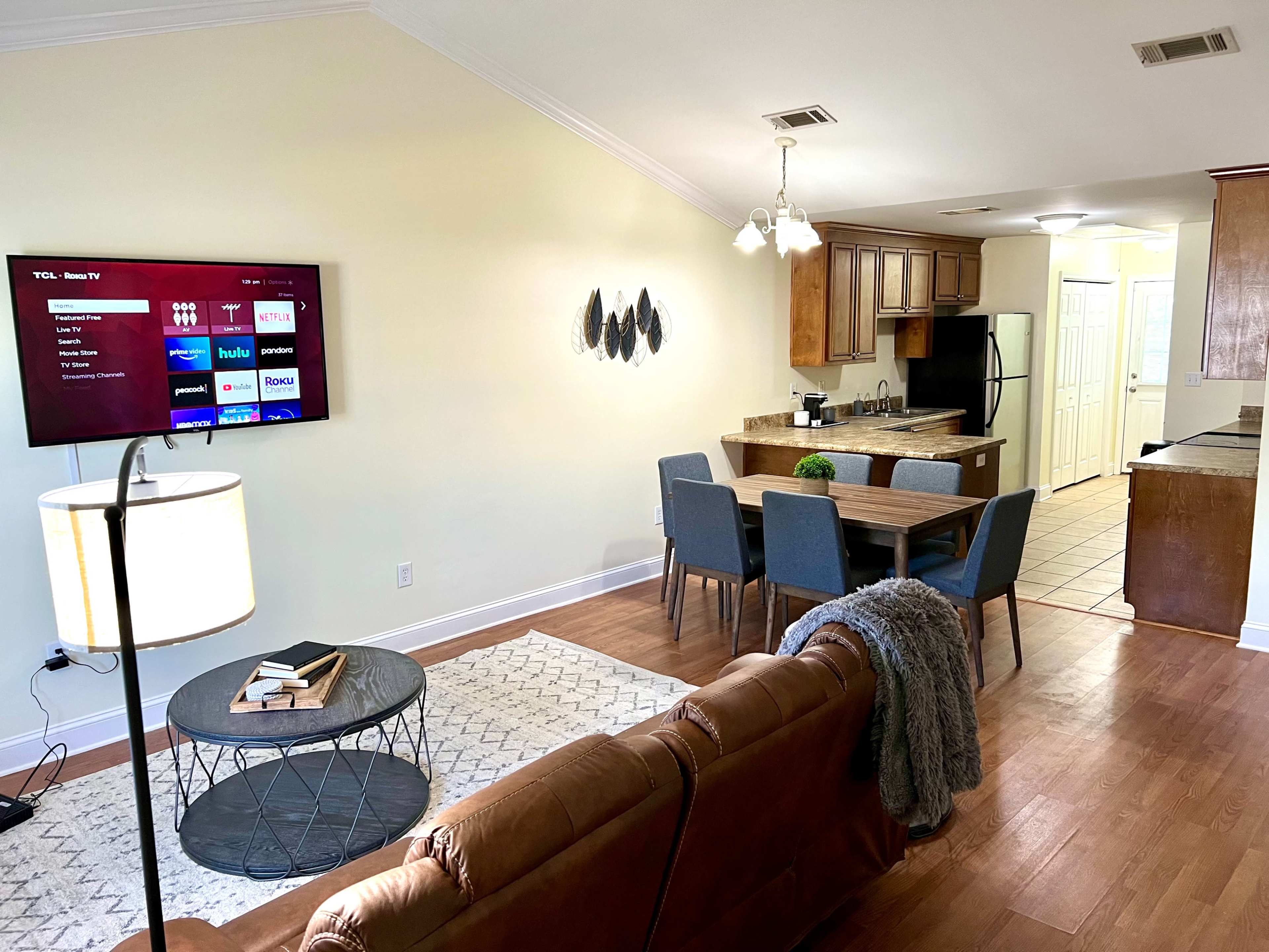 A living area with a brown leather sofa, a round coffee table, a television mounted on the wall, a dining table with chairs, and a kitchen visible in the background.