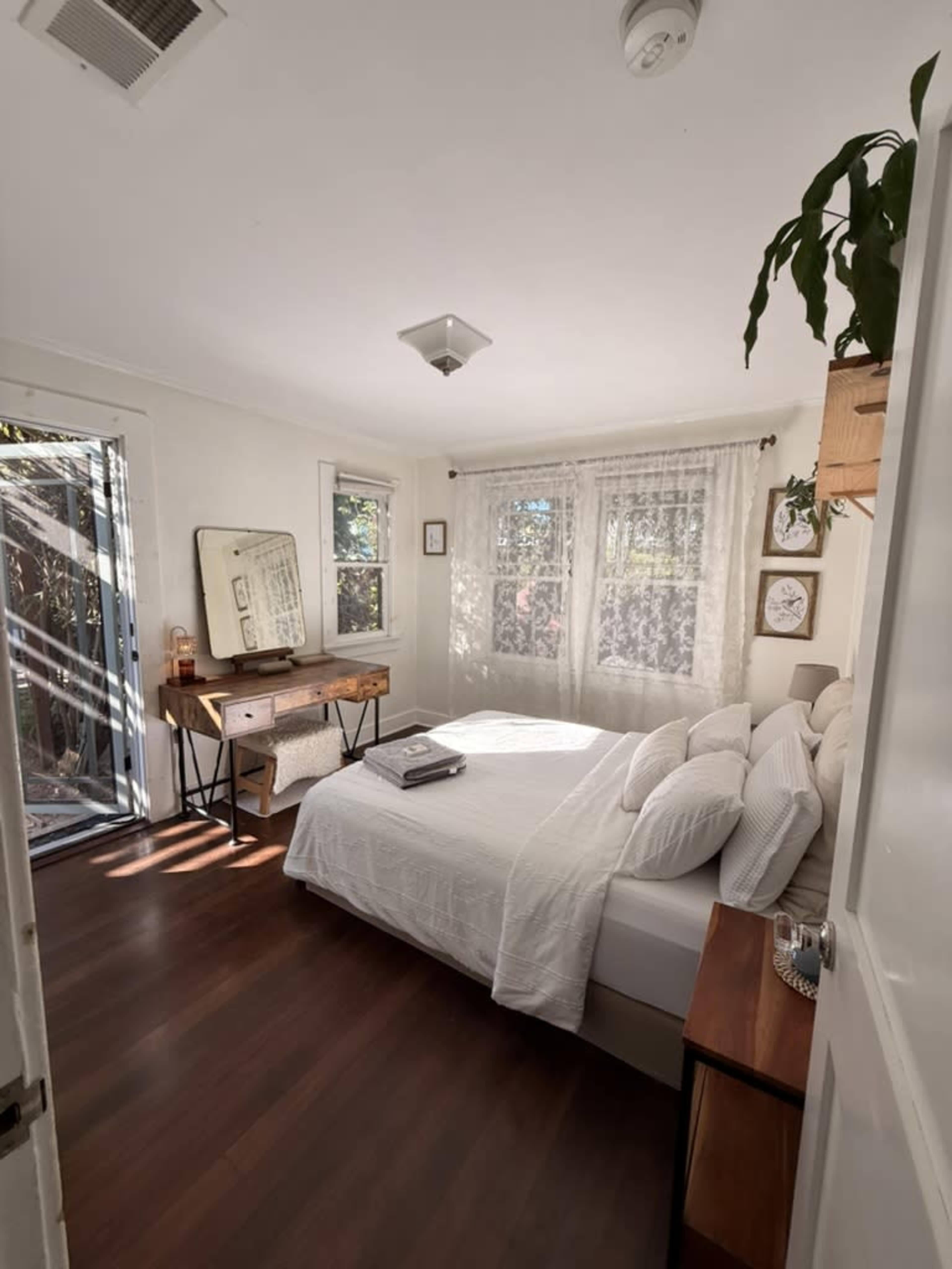 The image shows a cozy bedroom with a bed, a wooden desk, and large windows allowing natural light to enter.