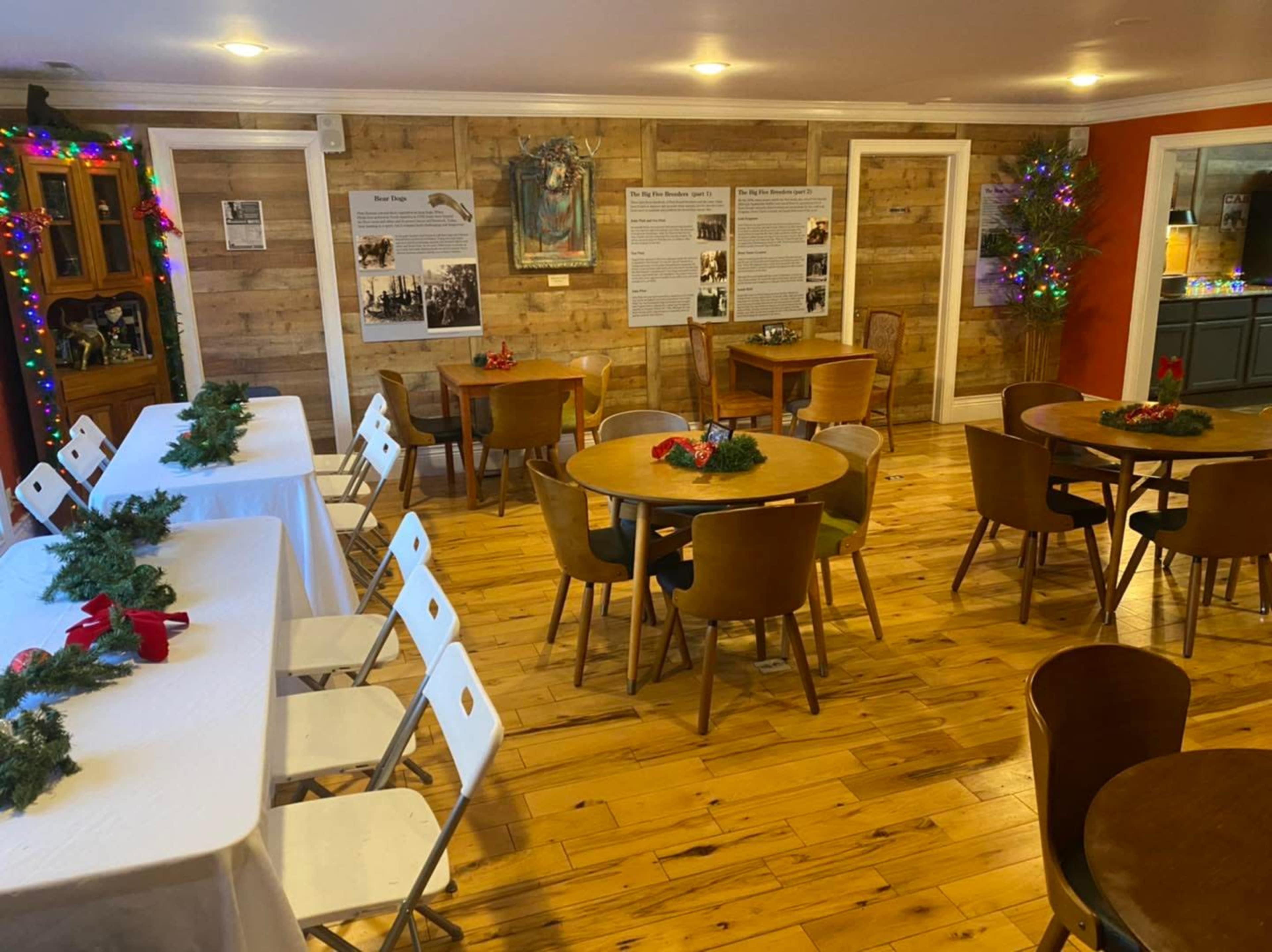 The room features wooden floors and walls with several round tables set for a gathering, decorated with greenery and surrounded by festive lights.