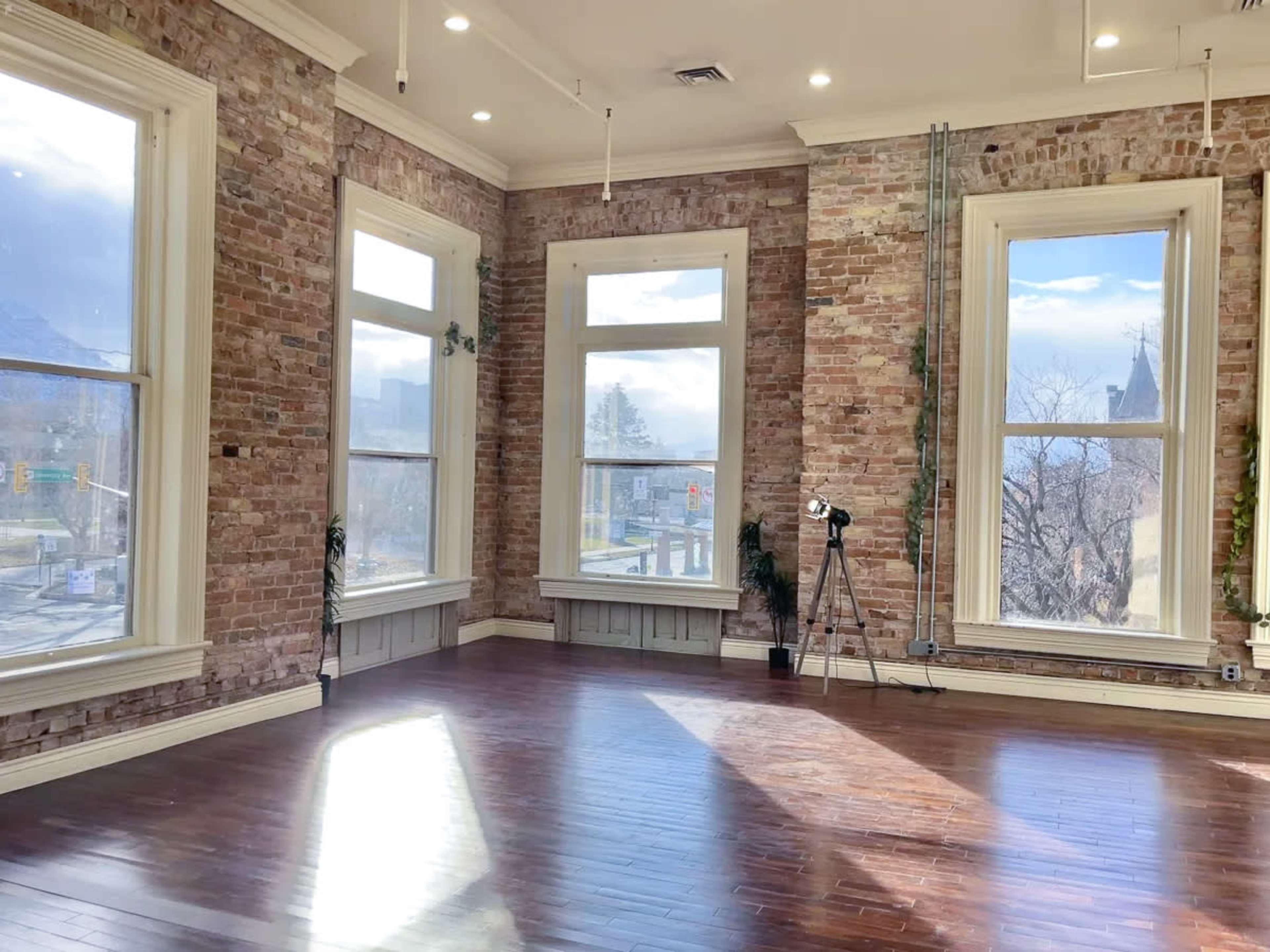 An empty room with exposed brick walls, large windows, and wooden flooring.