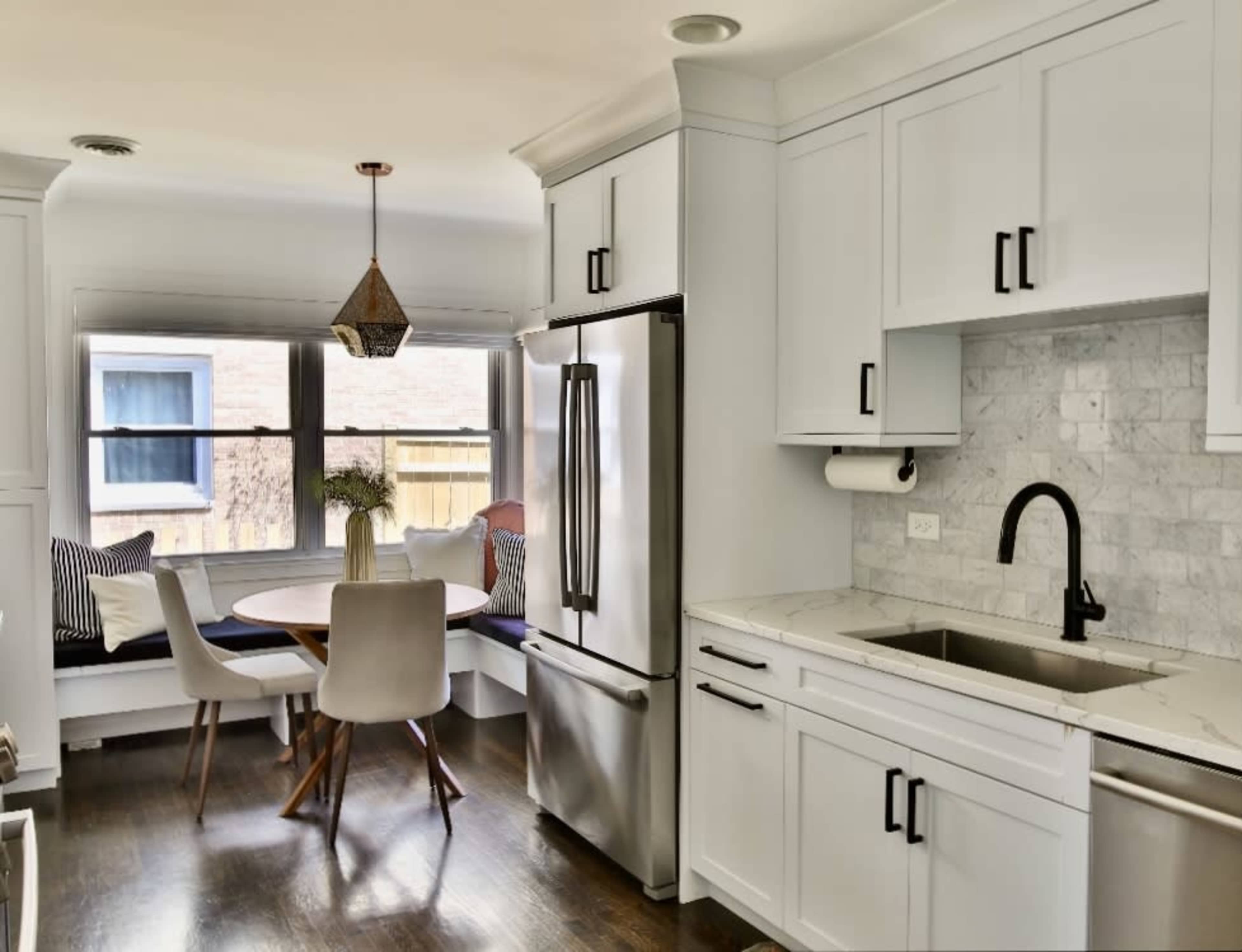 A modern kitchen features a stainless steel refrigerator, a marble countertop, a sink, and a circular dining table surrounded by chairs near a window bench.