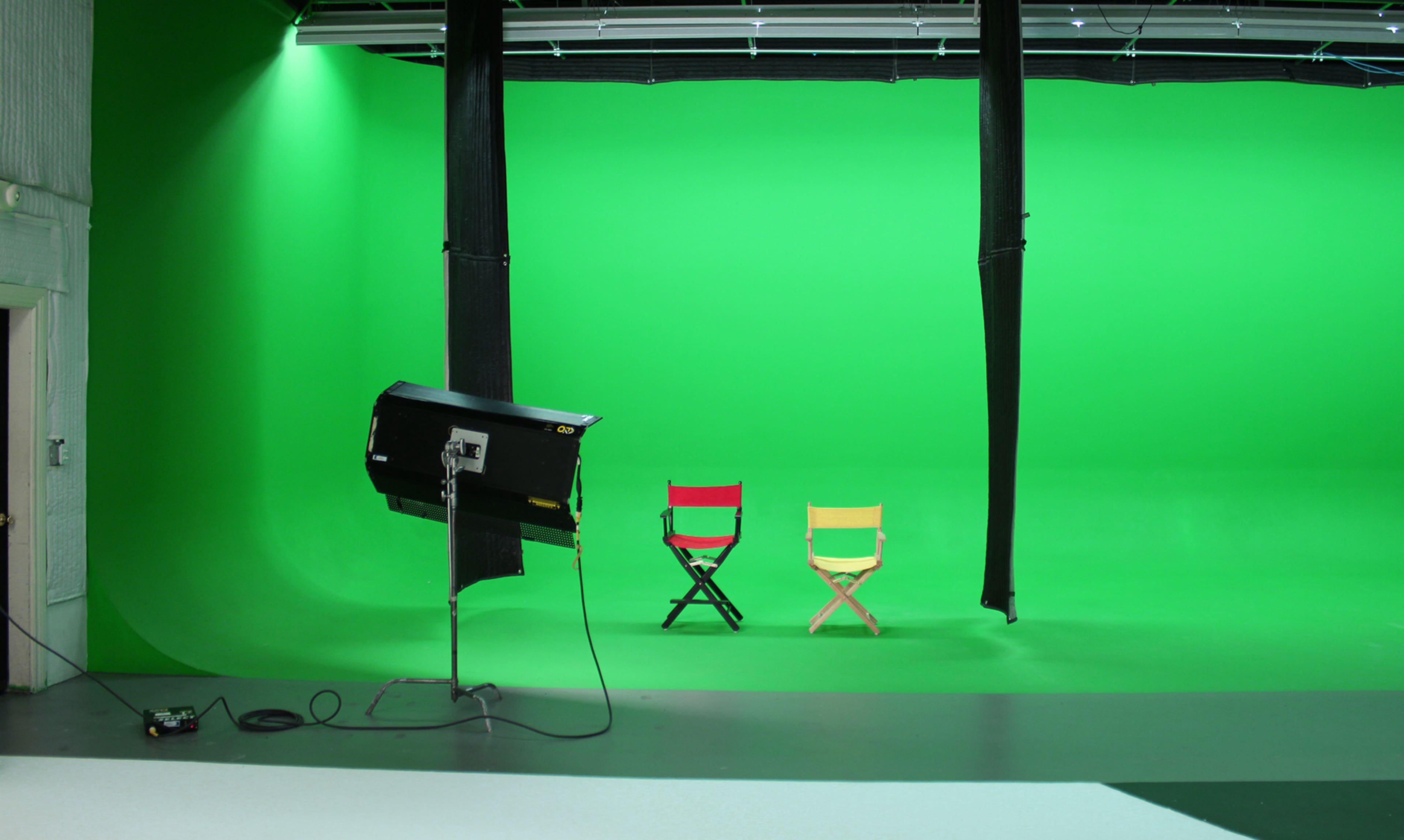 A photography studio with a green screen backdrop and two empty directors' chairs, one red and one yellow, positioned in front.