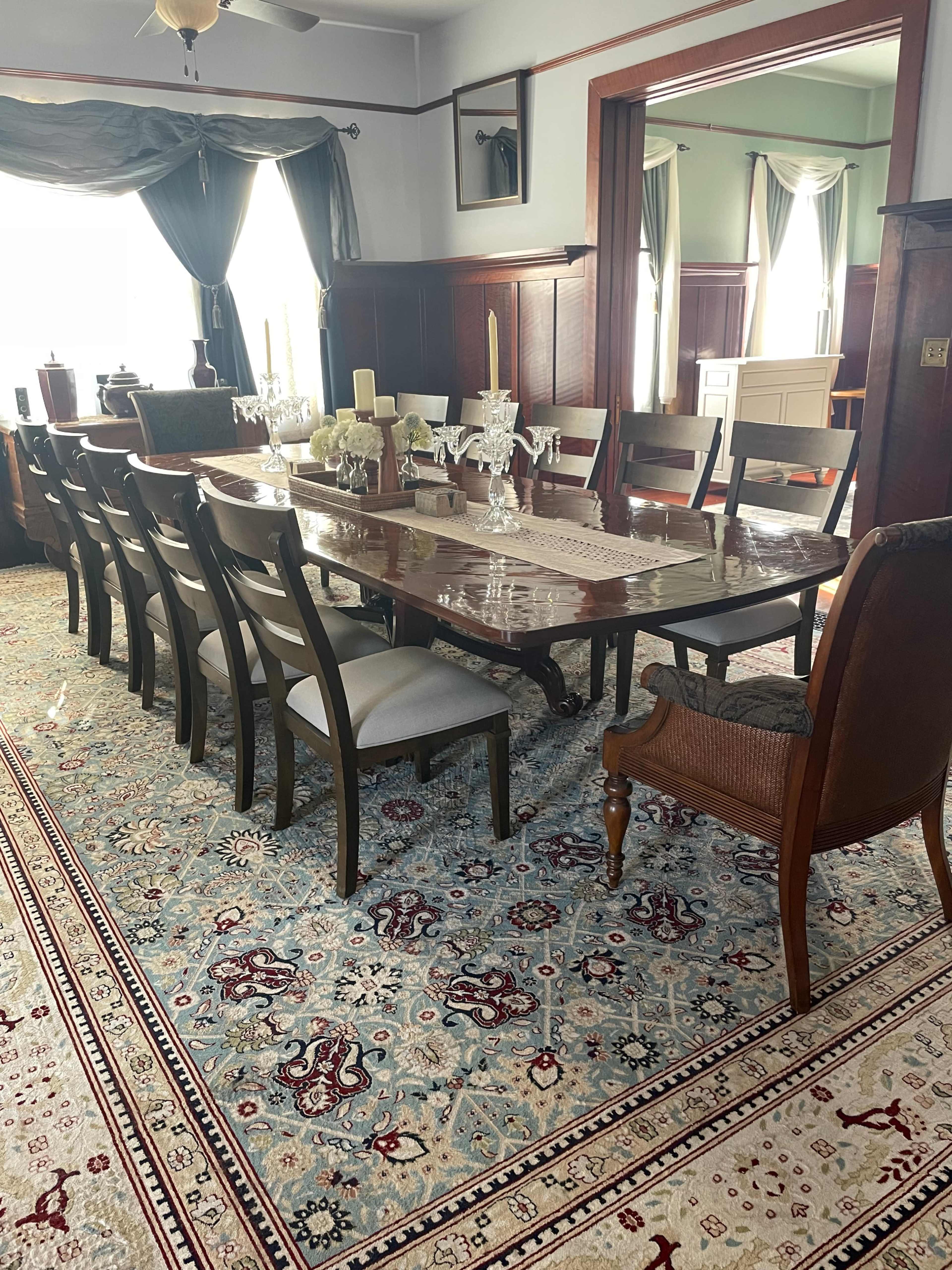 A spacious dining room with a long wooden table set for dinner, surrounded by chairs and featuring ornate rugs and curtains.