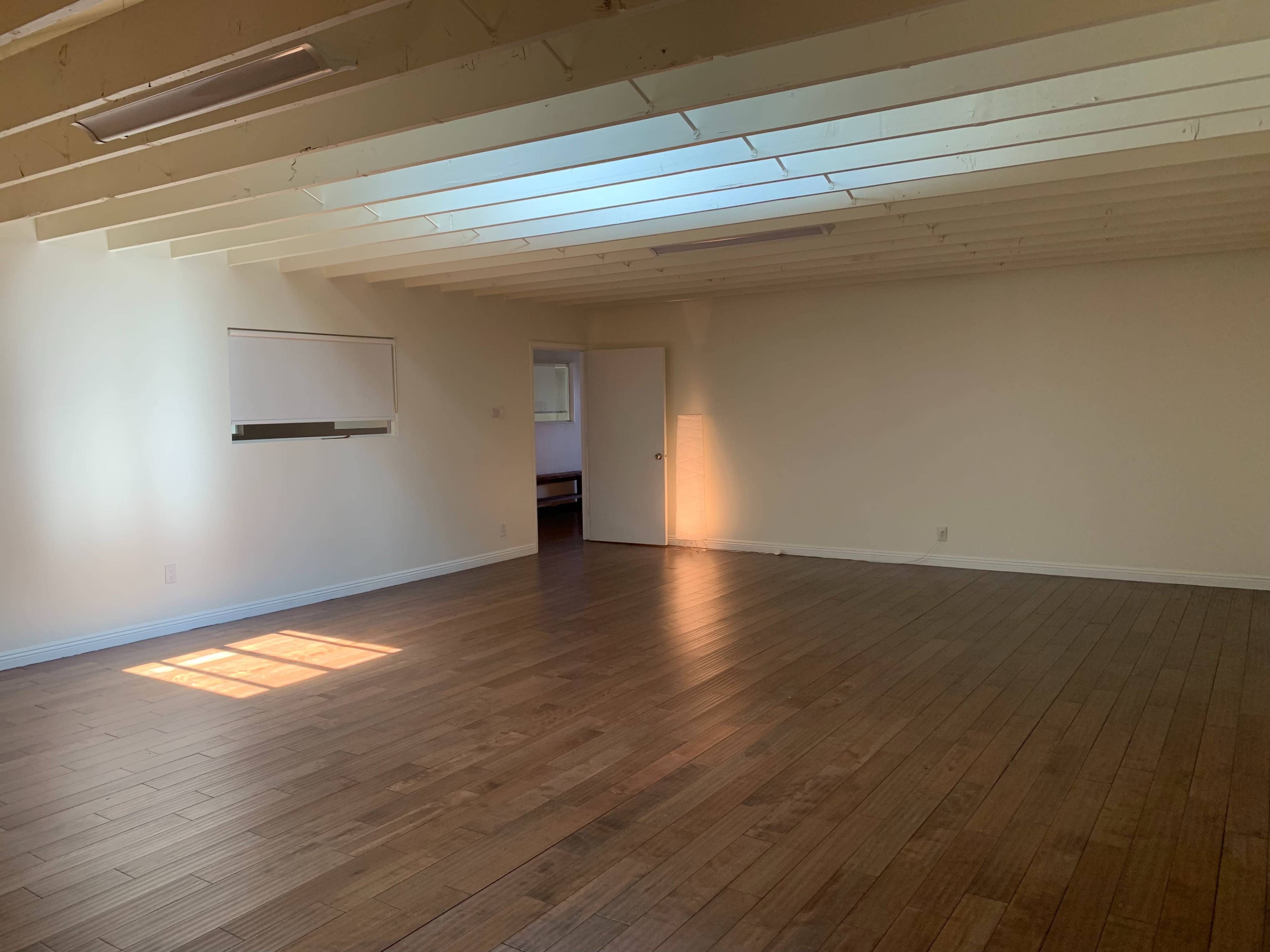 A spacious interior room with wooden flooring, white walls, and exposed beams on the ceiling, illuminated by natural light from a window.