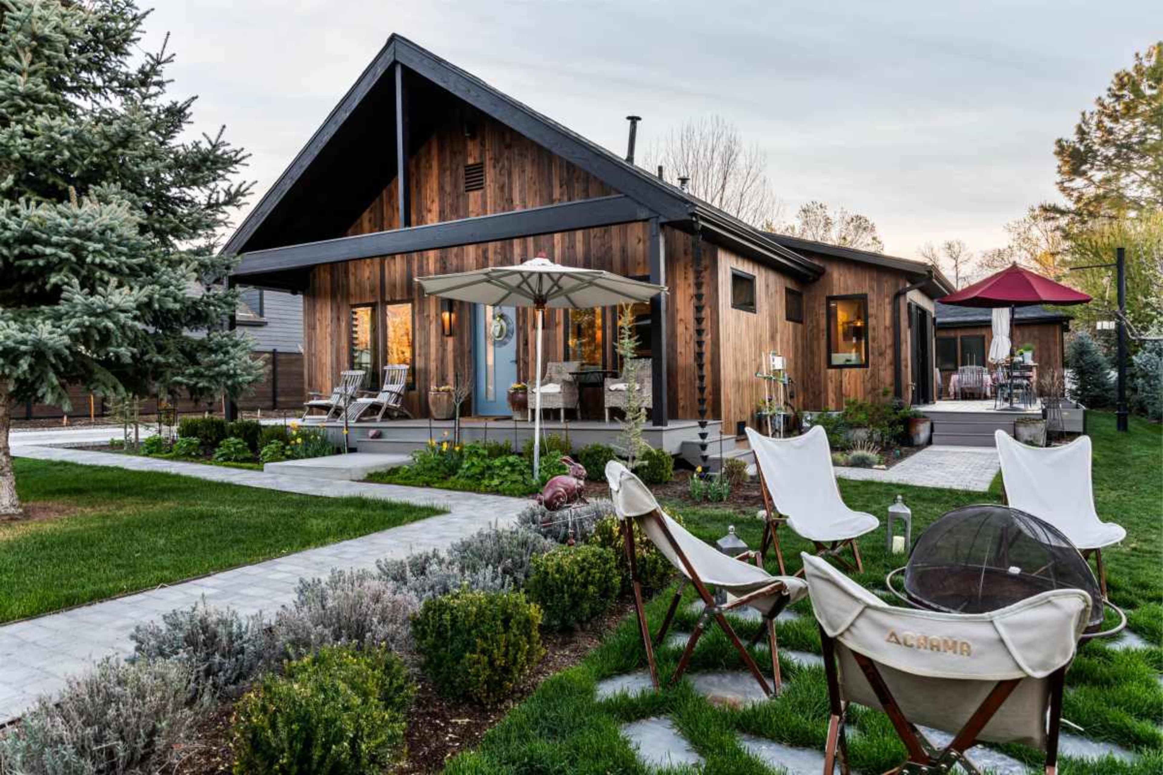 A modern wooden house with large windows is surrounded by landscaped gardens and outdoor seating areas.