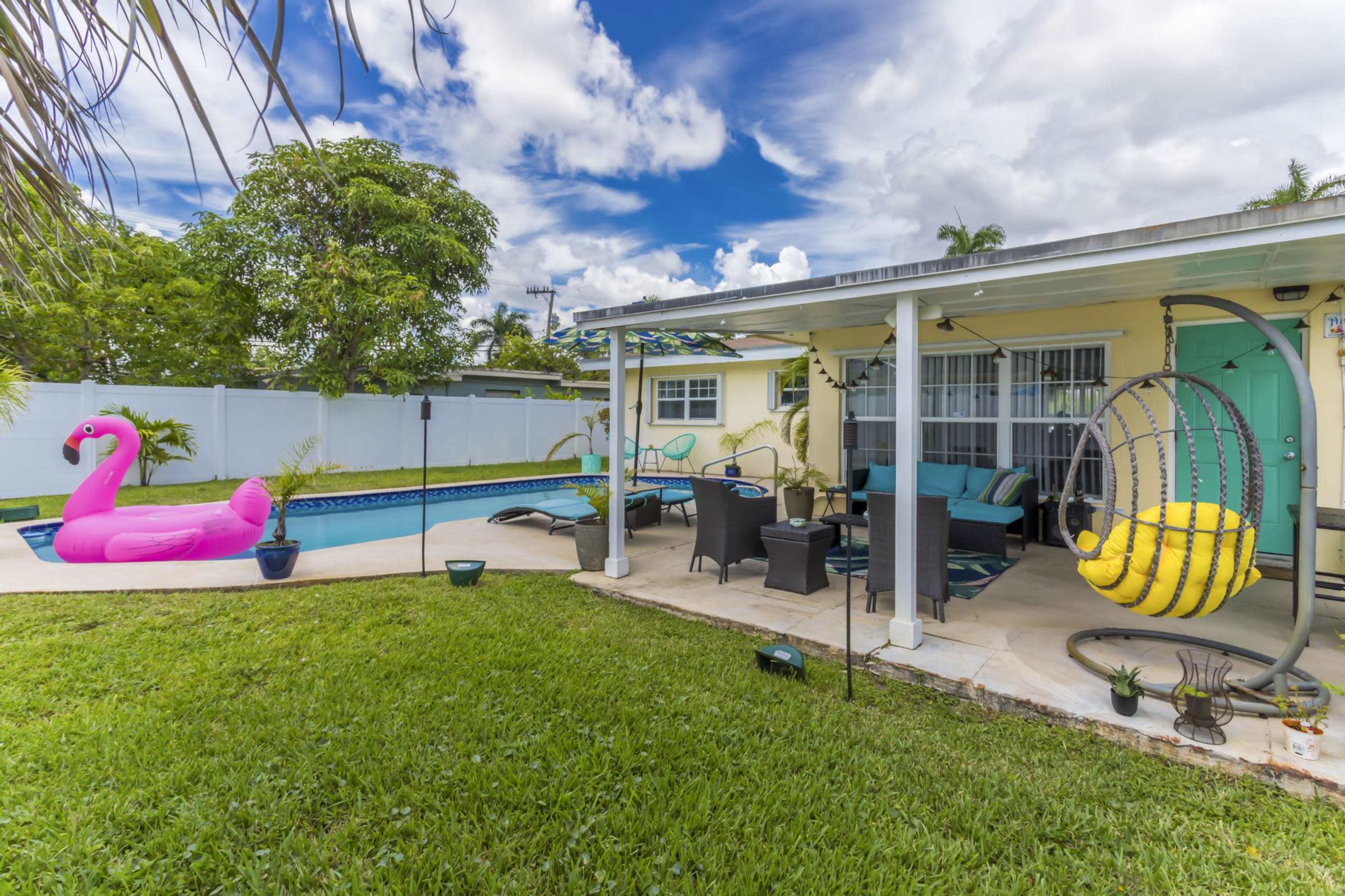 The image features a backyard pool area with a bright pink flamingo float, a hanging swing chair, and lounge furniture under a covered patio.