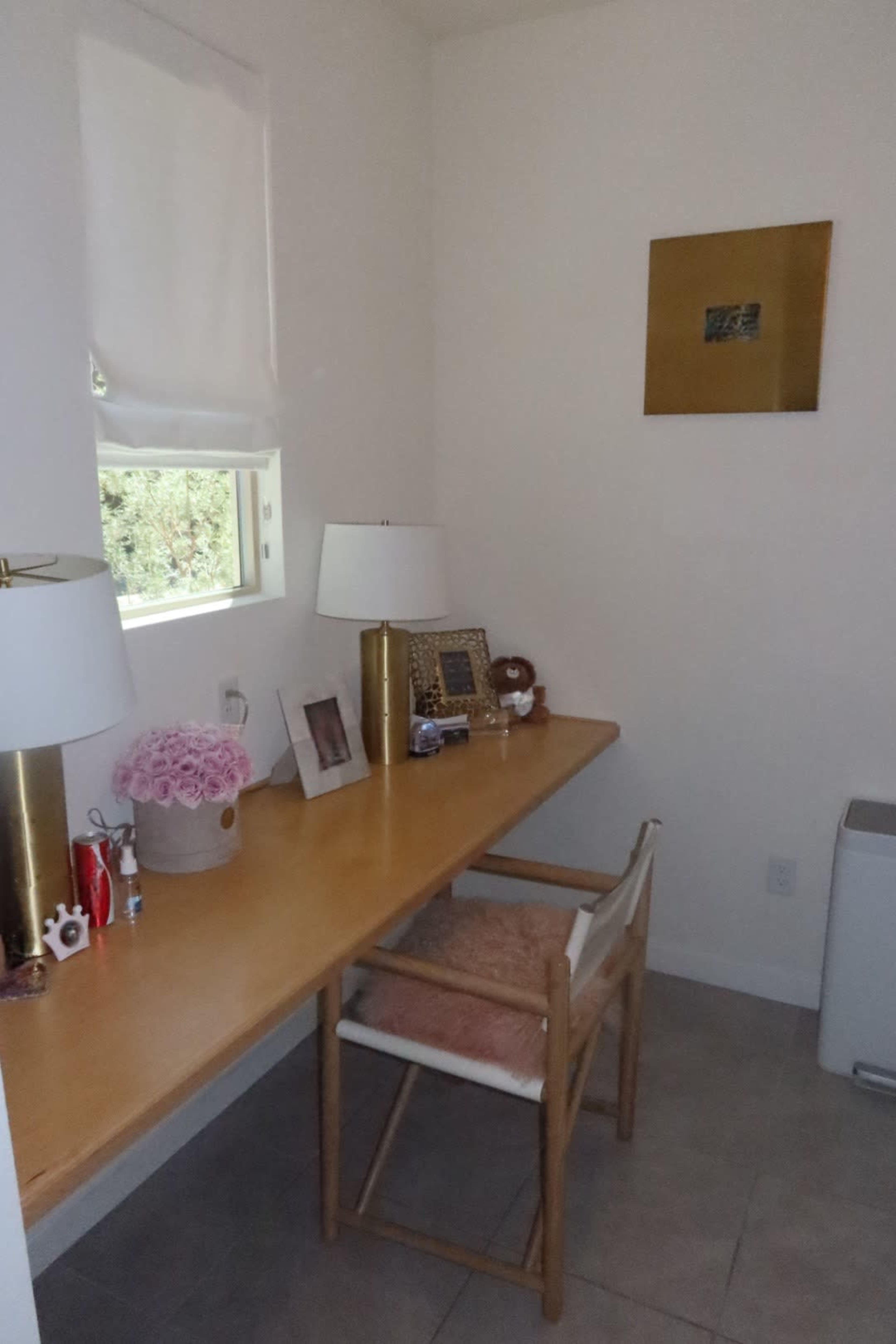 A small home office features a wooden desk with a chair, two lamps, decorative items, and a window illuminating the space.