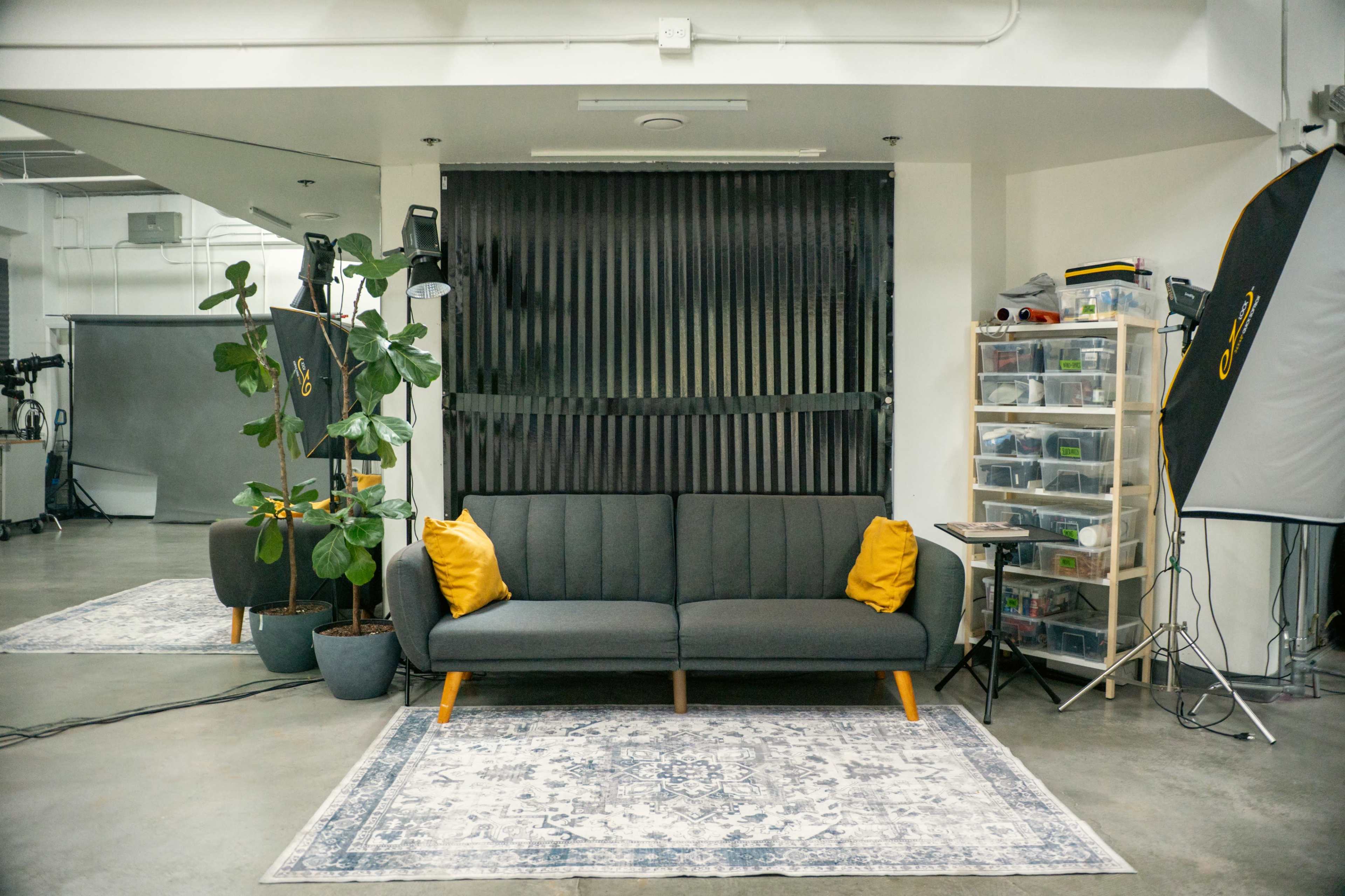 The image depicts a minimalist studio space featuring a gray sofa with yellow pillows, a tall plant, a shelving unit with transparent storage bins, and professional lighting equipment.