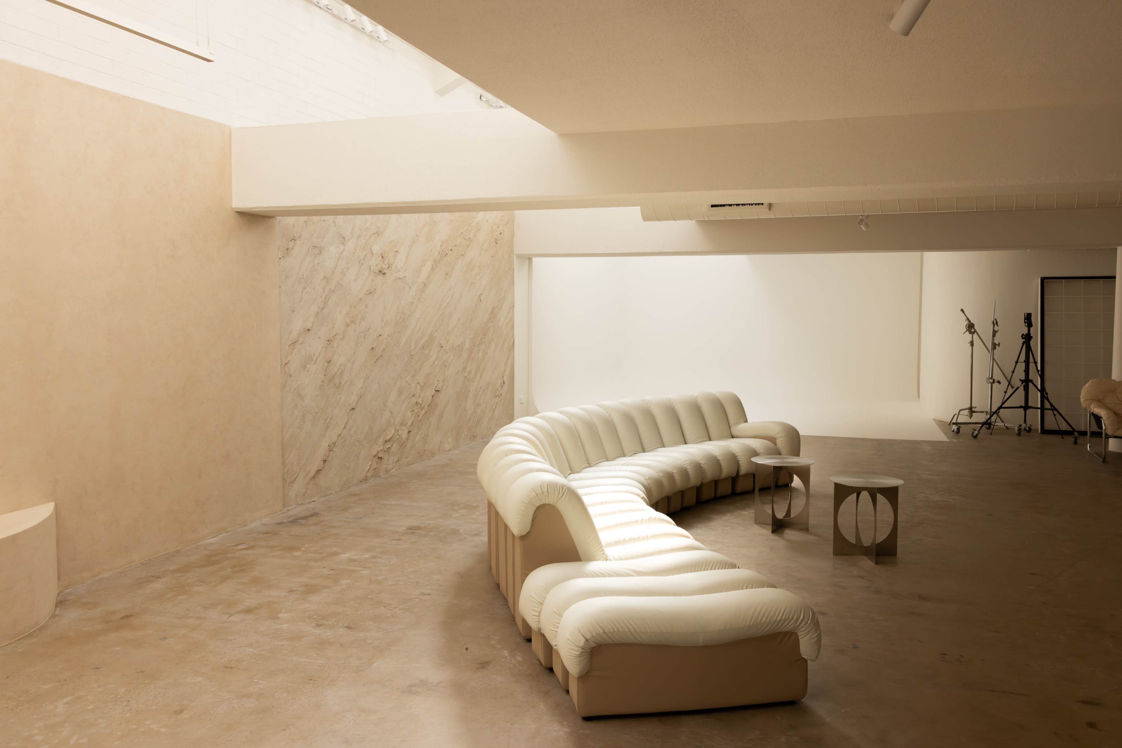 A curved white sofa is positioned in a spacious, minimalist room with beige walls and a simple coffee table nearby.