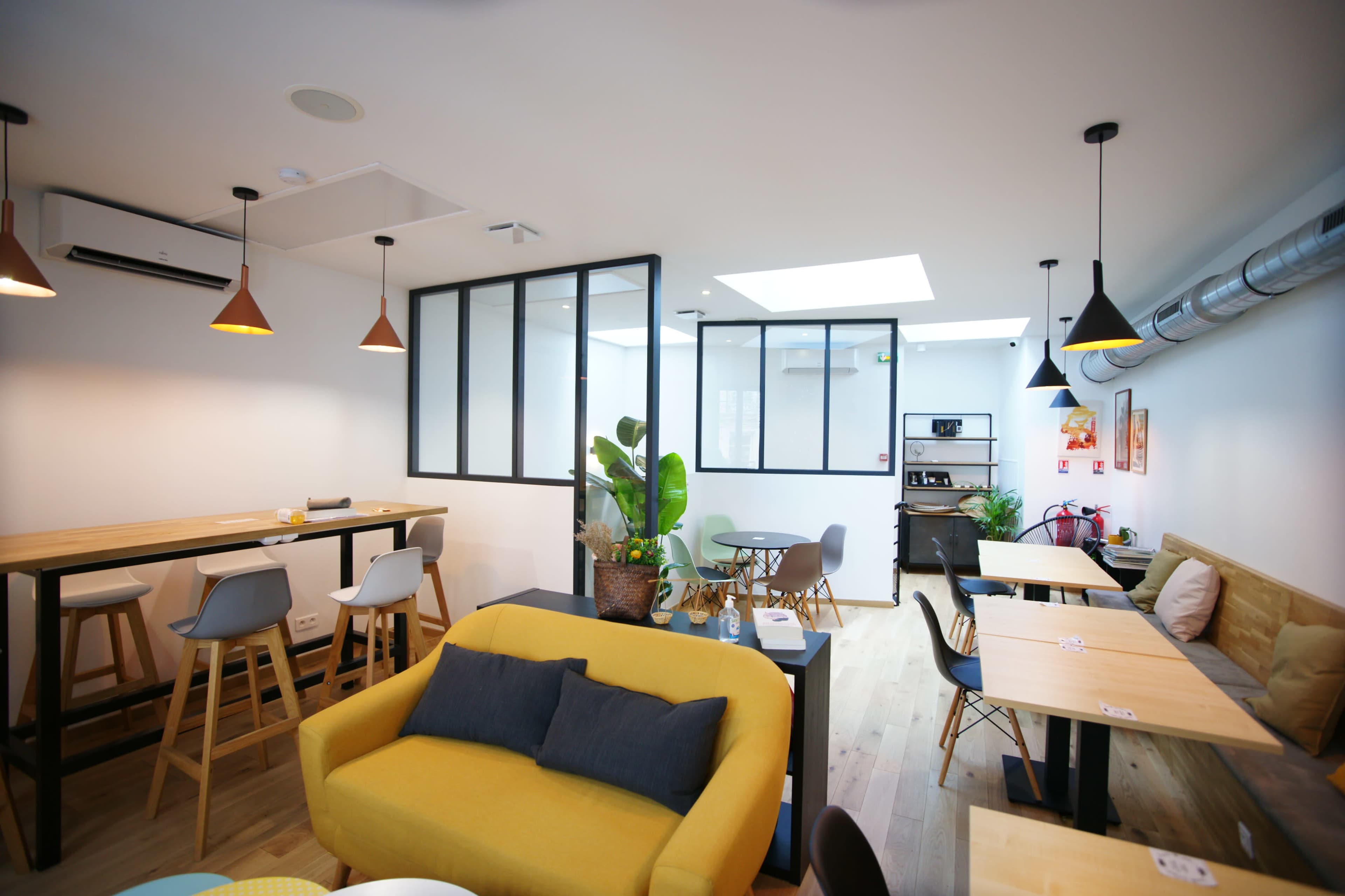 A modern café interior with wooden tables, a yellow sofa, and large windows allowing natural light to fill the space.