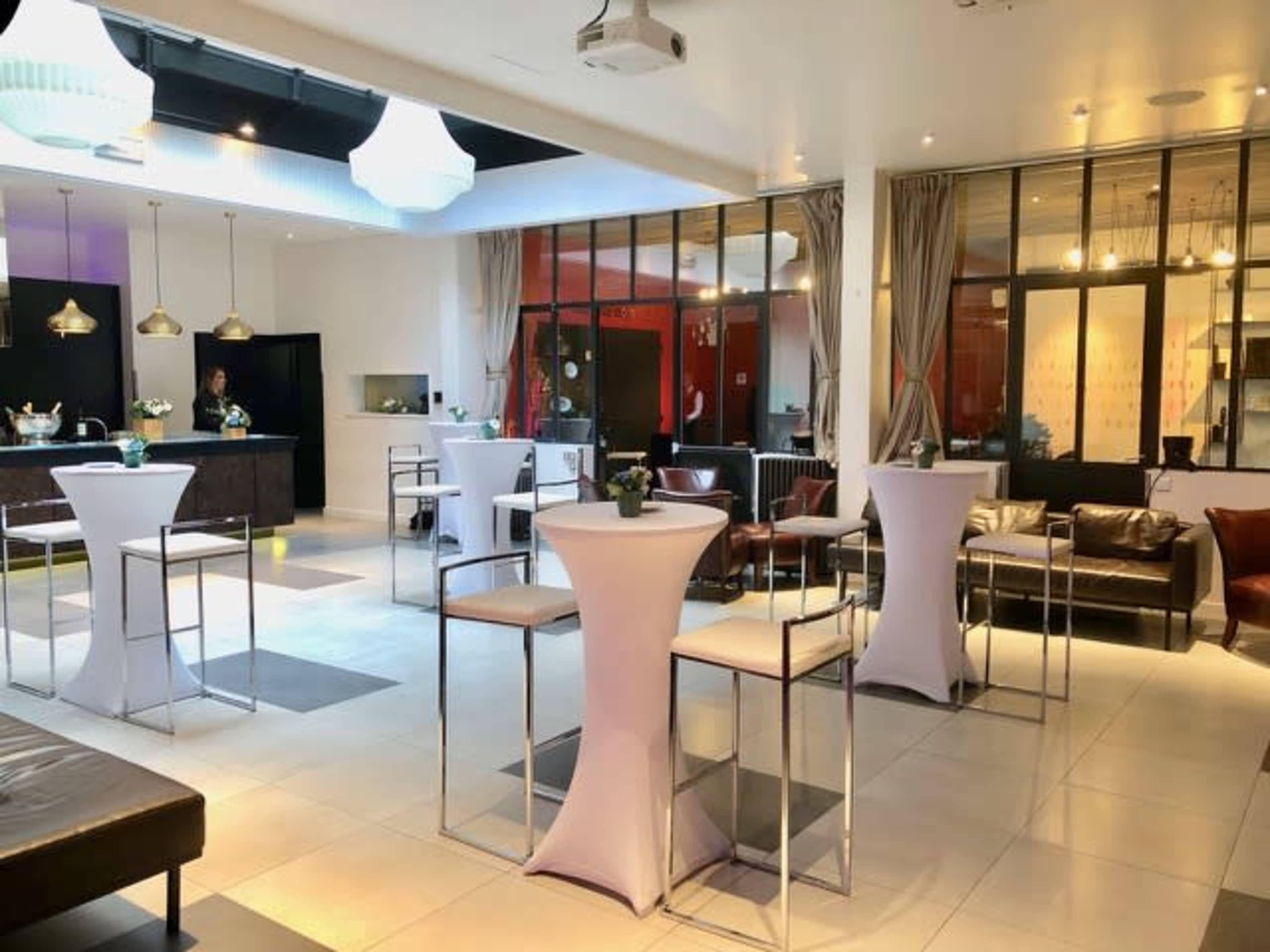 A modern event space with high tables and chairs, a bar area, and large windows providing a view of a lounge area.