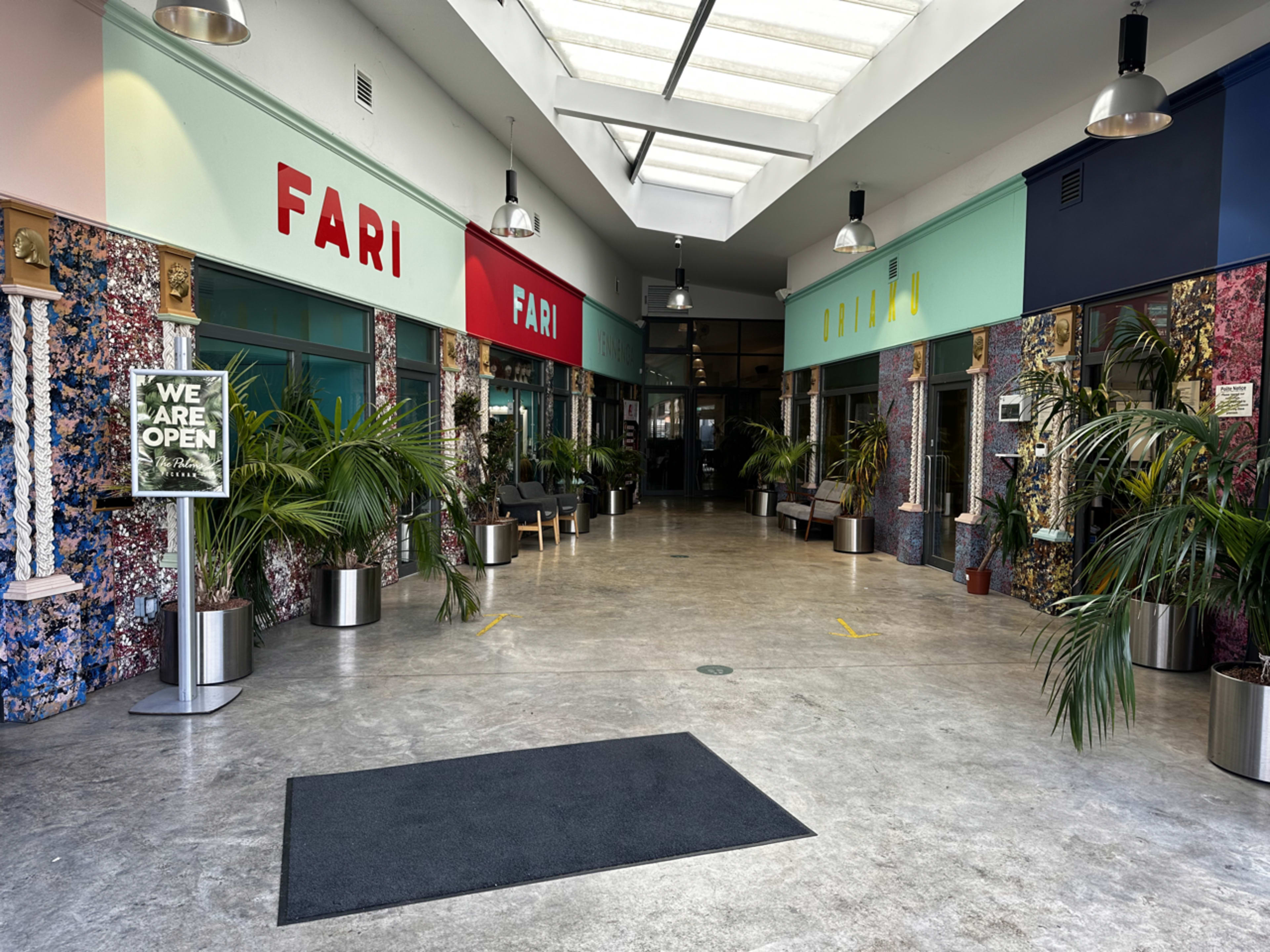 The image shows a brightly lit indoor corridor with potted plants and colorful signage, indicating various businesses.