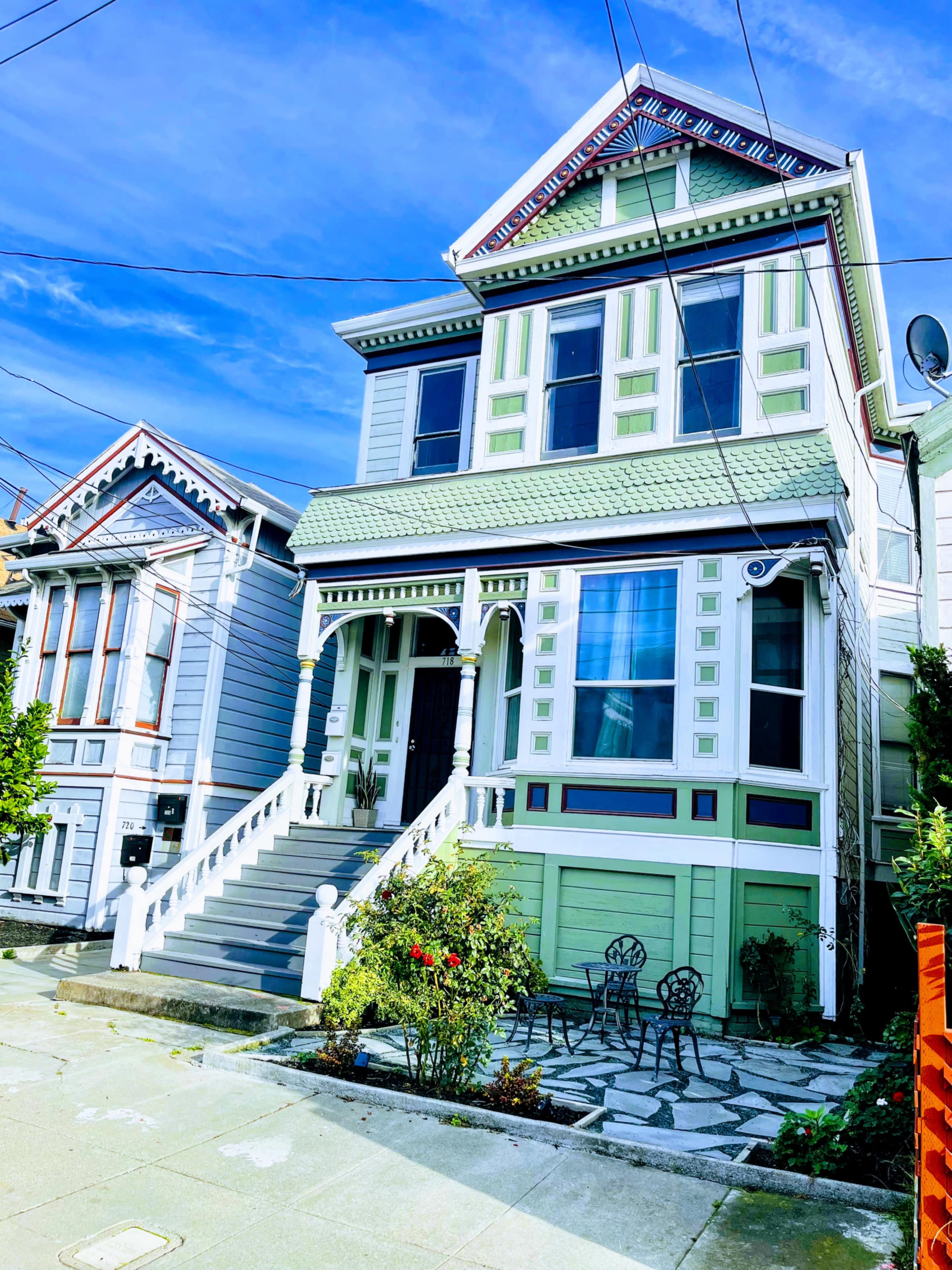 A Victorian-style house with green and white trim, featuring a front porch and decorative details, stands on a residential street.