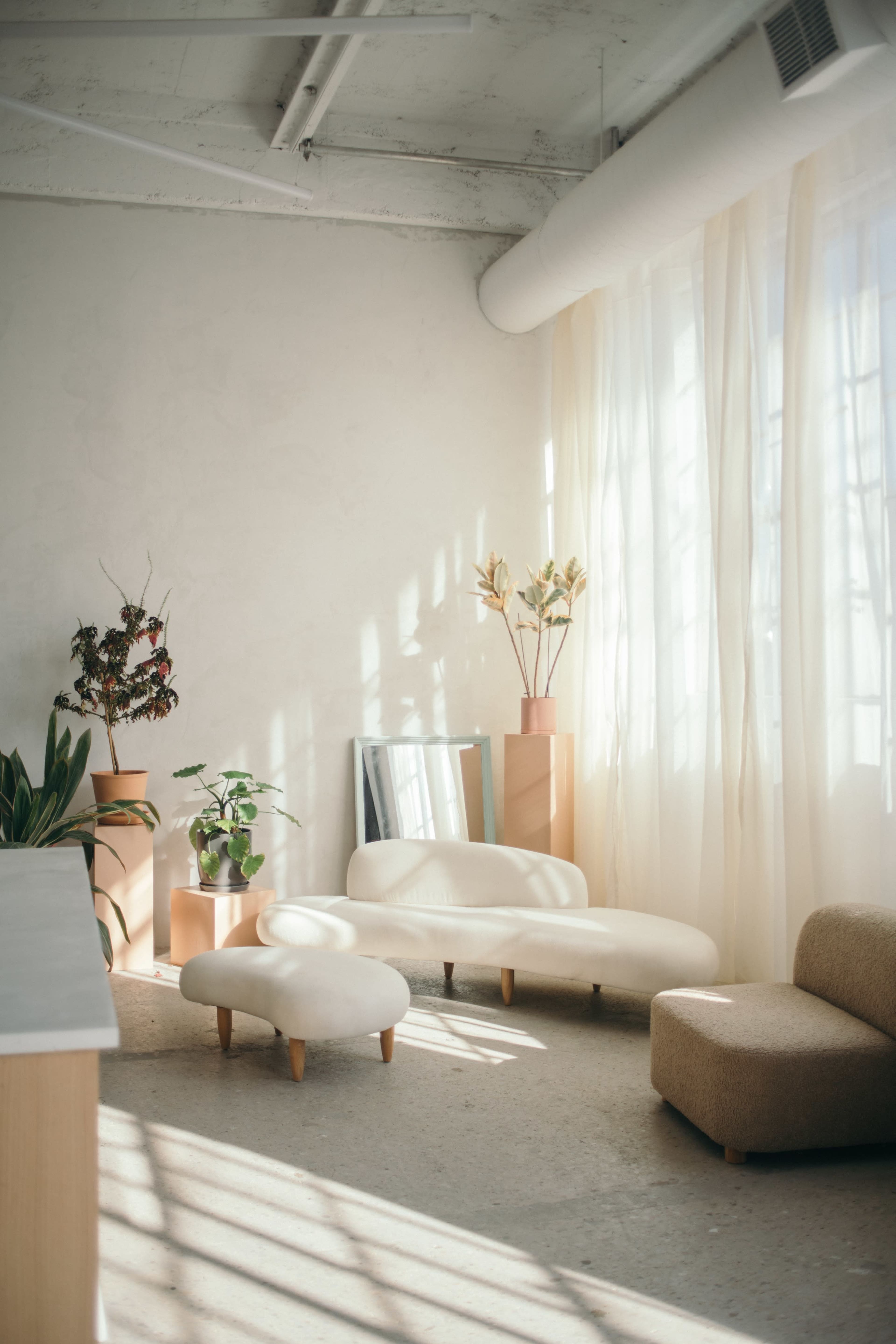 A bright interior features light-colored furniture, plants, and decorative elements arranged near large windows with flowing curtains.