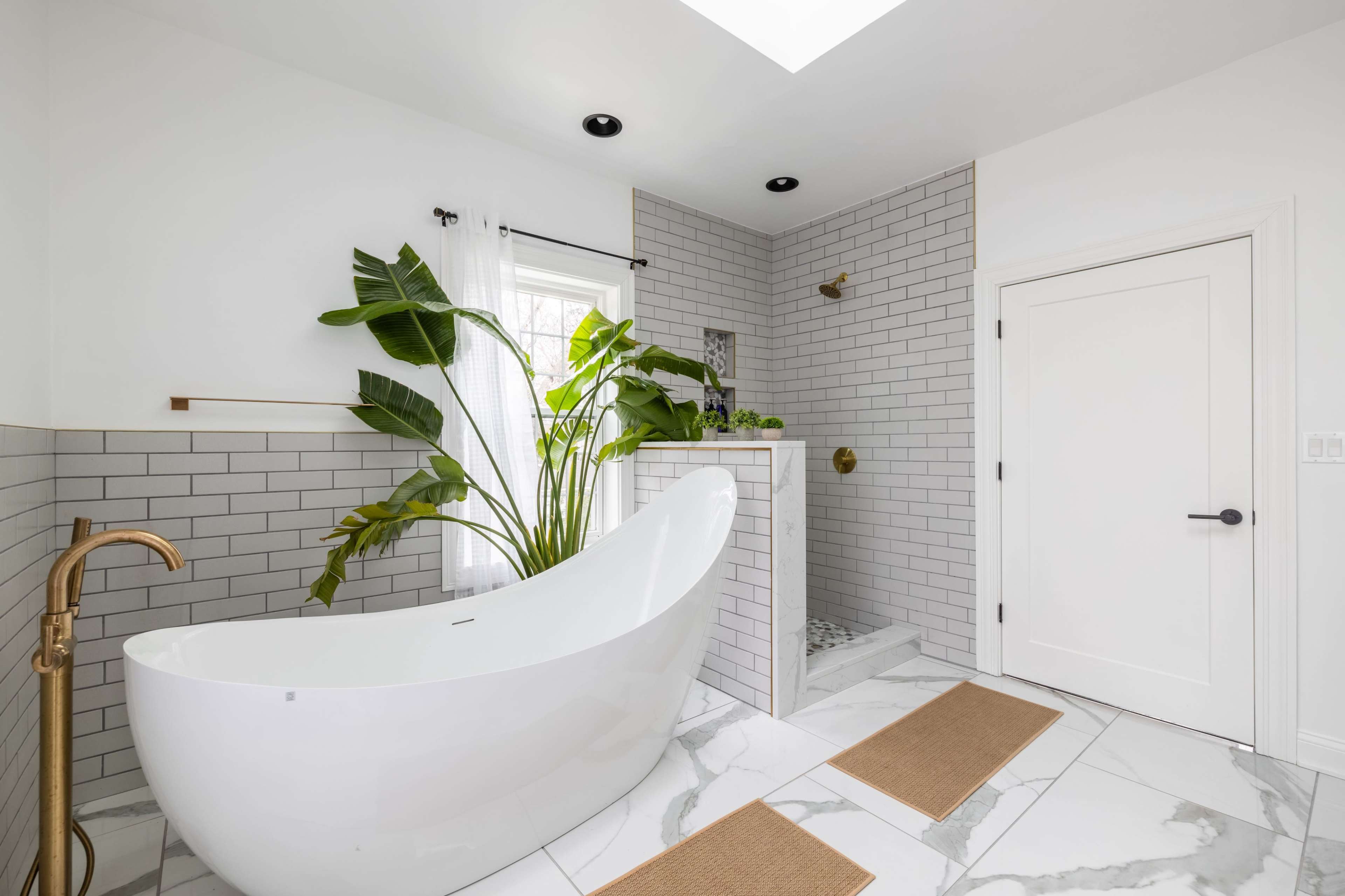 The bathroom features a freestanding white bathtub, a glass-enclosed shower, and large potted plants set against a backdrop of white tile and marble flooring.