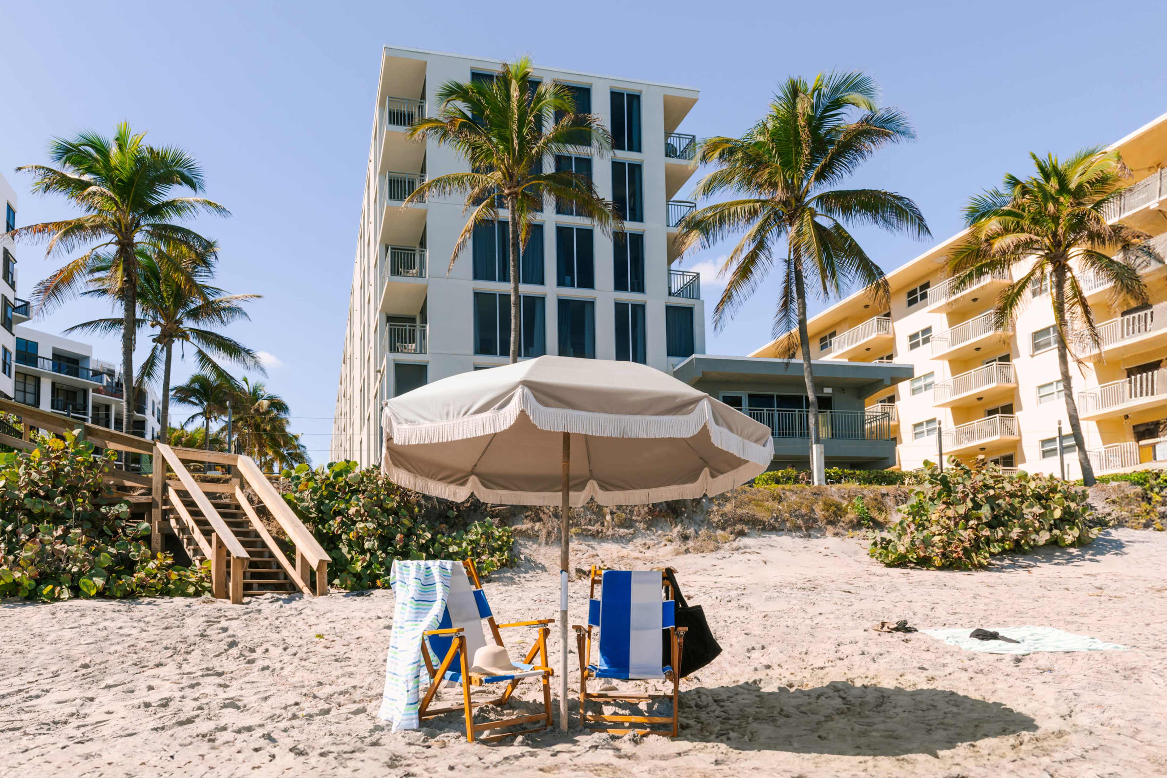 Two beach chairs and an umbrella are set up on the sand in front of a large hotel surrounded by palm trees.
