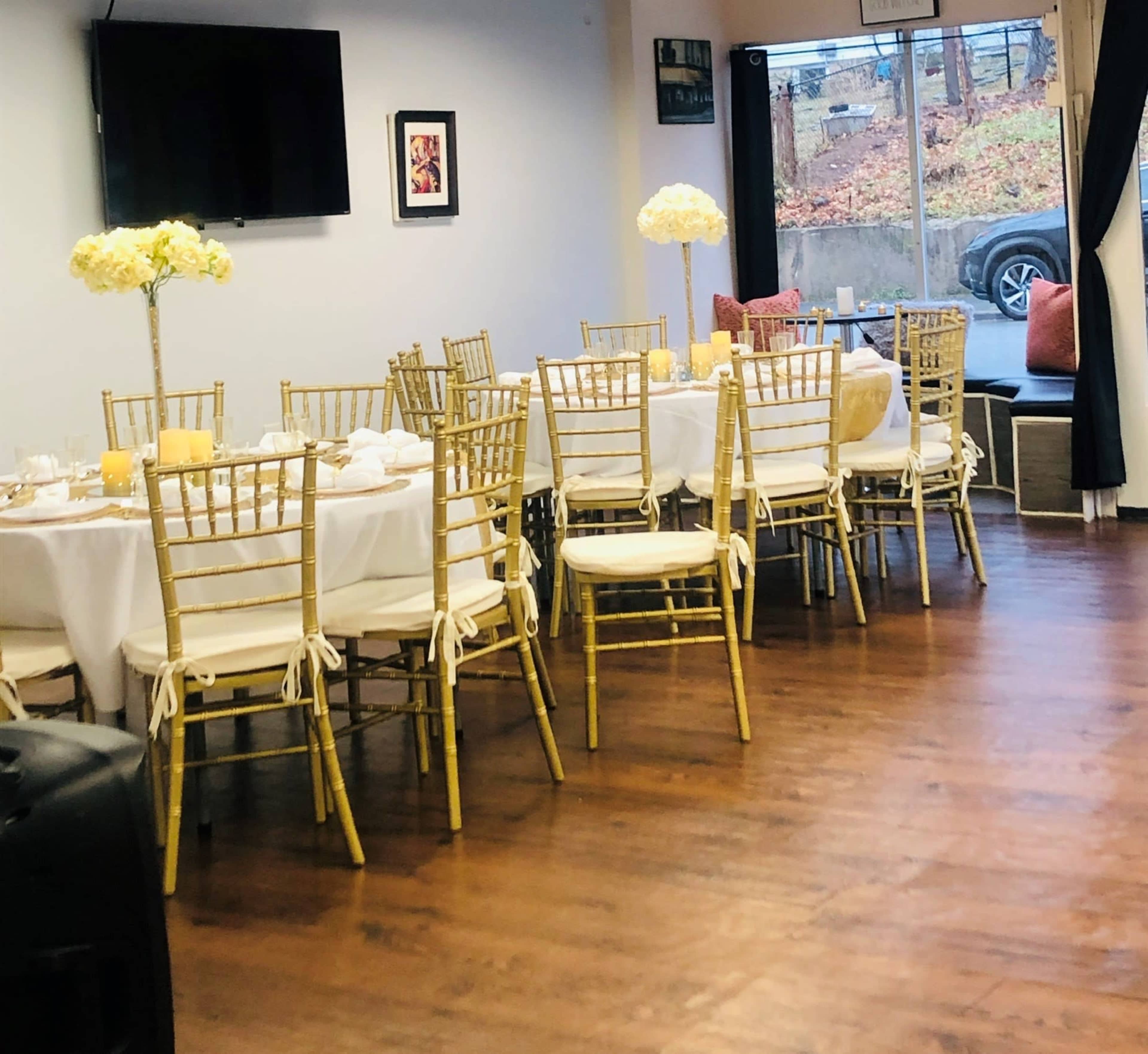 A dining area with gold chiavari chairs and tables set for a meal, featuring floral centerpieces and large windows overlooking an outdoor scene.