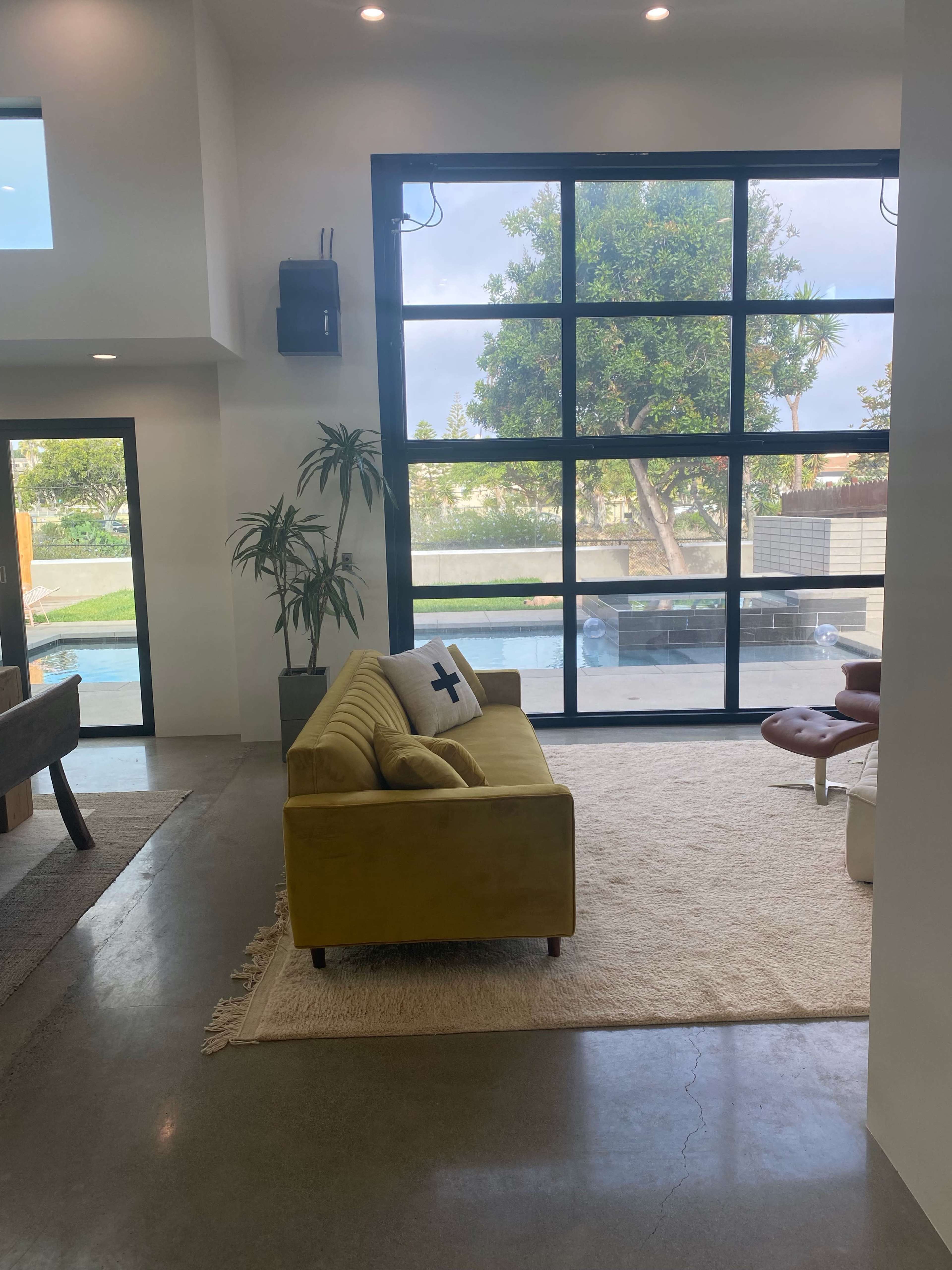 A modern living room features a yellow sofa positioned on a light-colored rug, with large windows allowing natural light and a view of greenery outside.